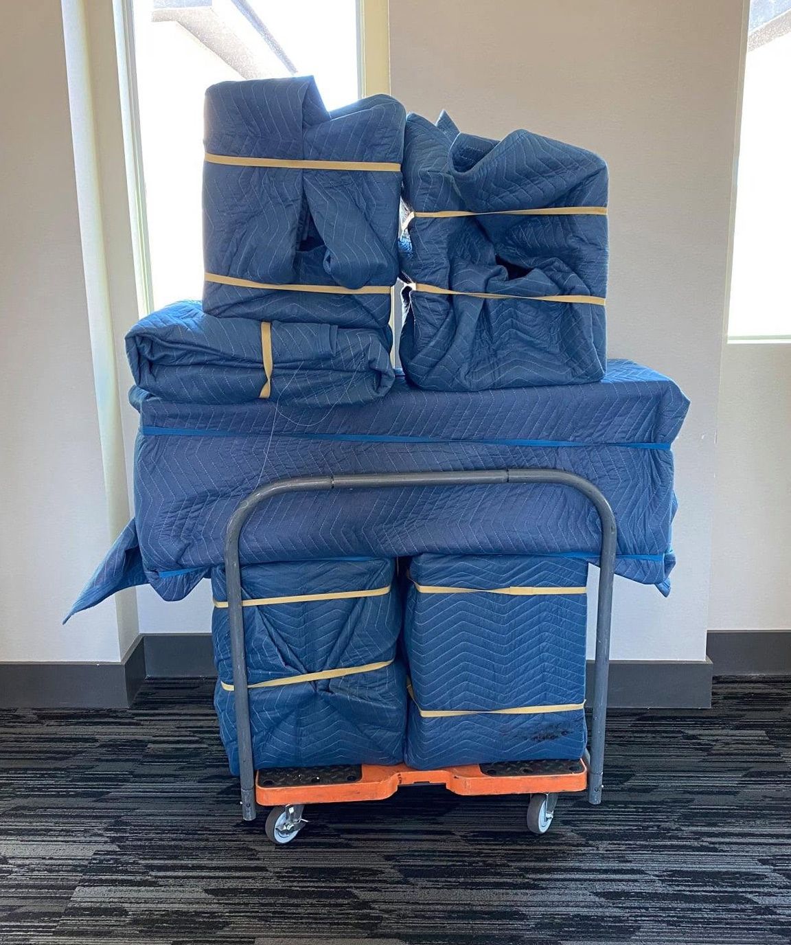 Stacked blue wrapped packages on a cart in a hallway near a window.