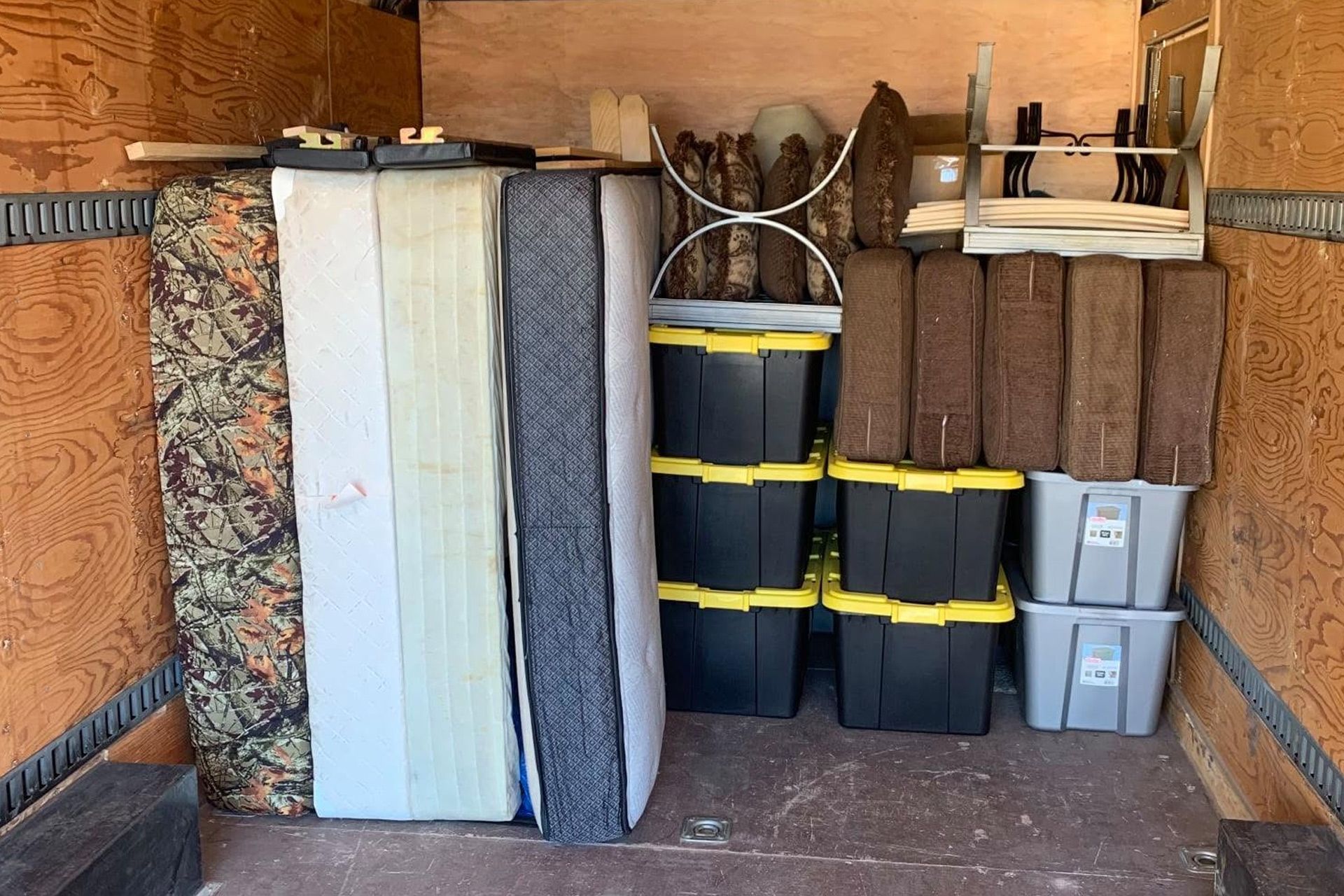 Storage trailer with stacked mattresses, rolled rugs, and plastic bins inside a wooden interior