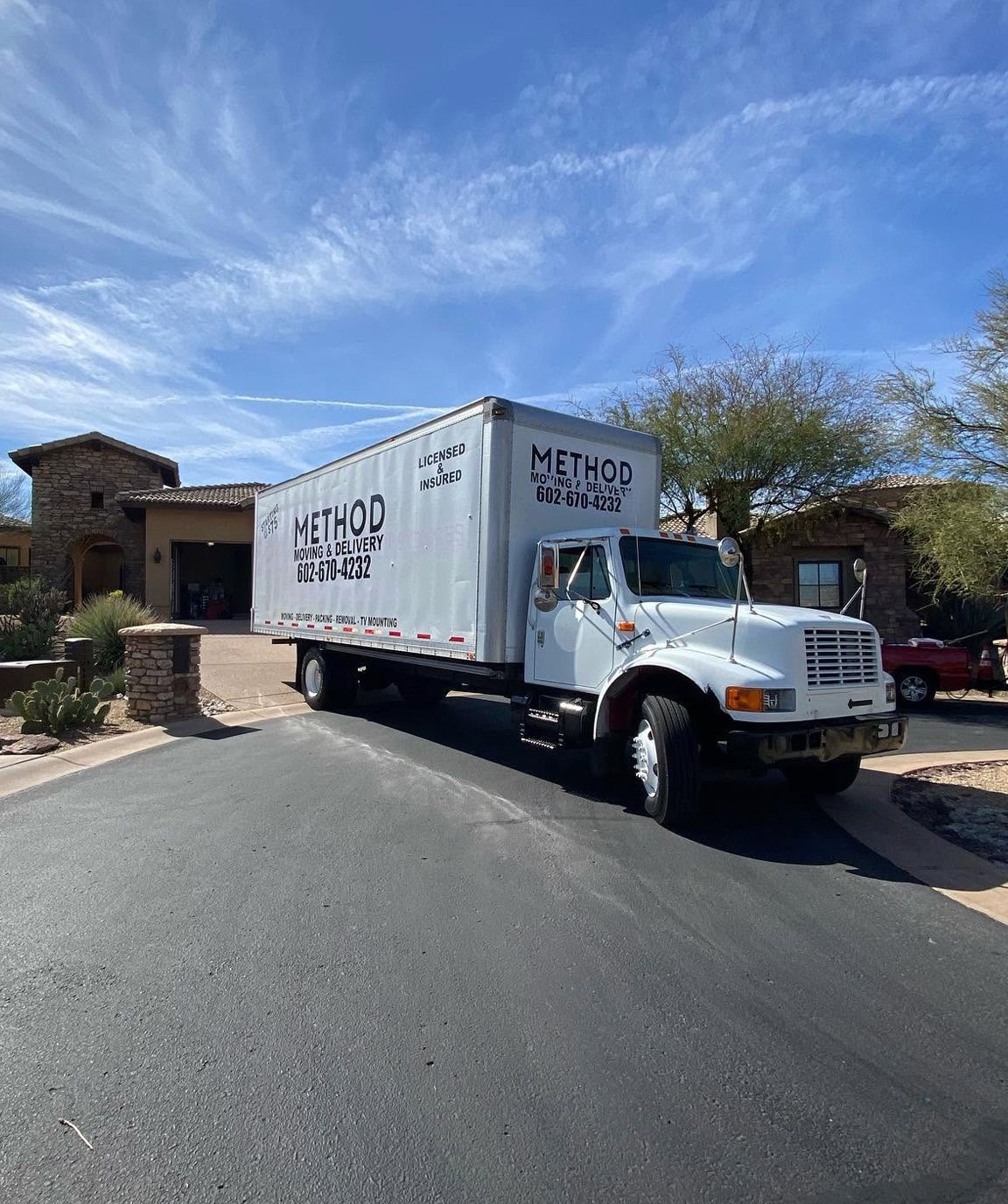 White moving truck on a suburban driveway under a blue sky, beside a house and brick wall