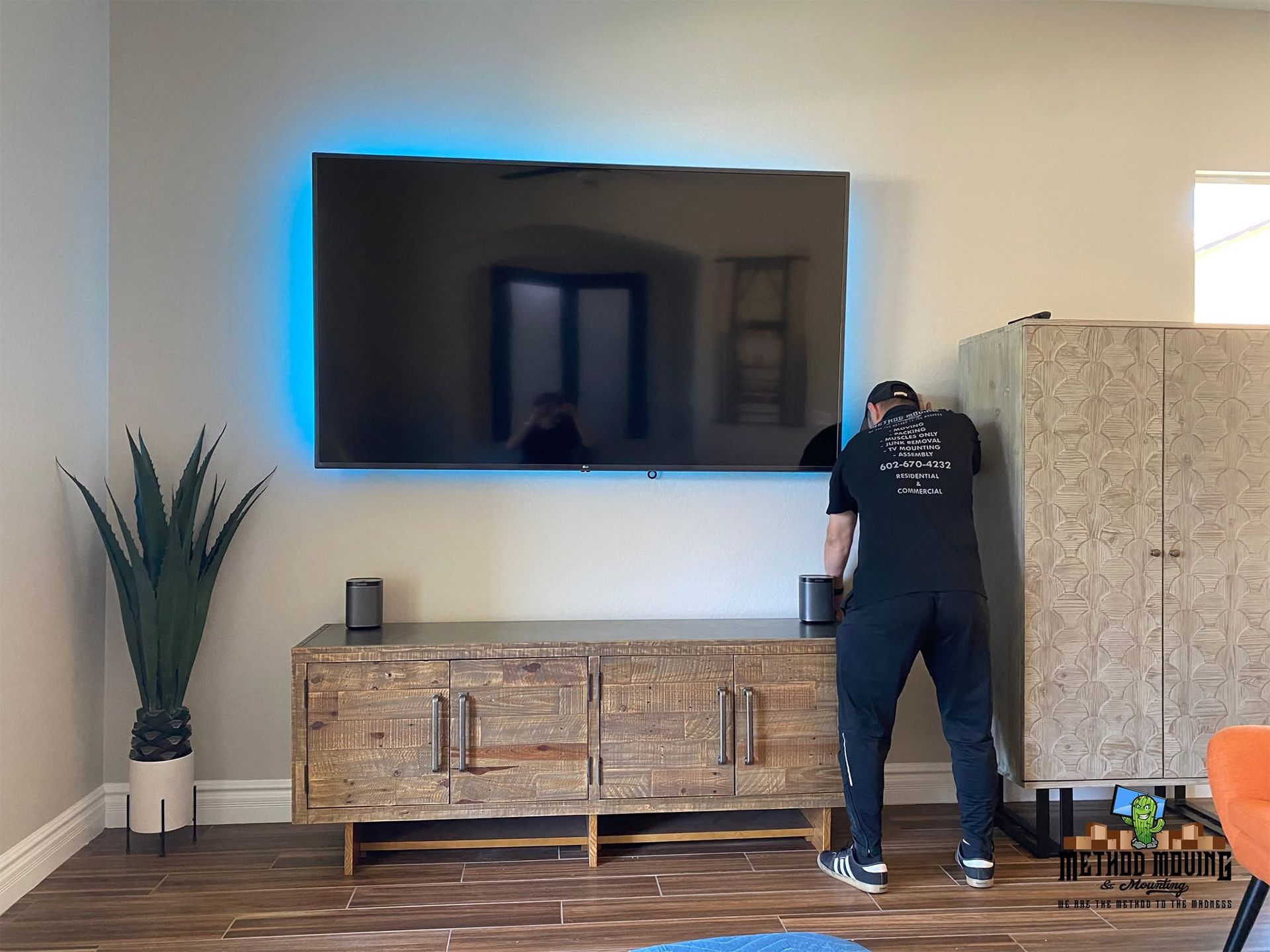 Person installing a wall-mounted TV above a wooden media console in a bright living room