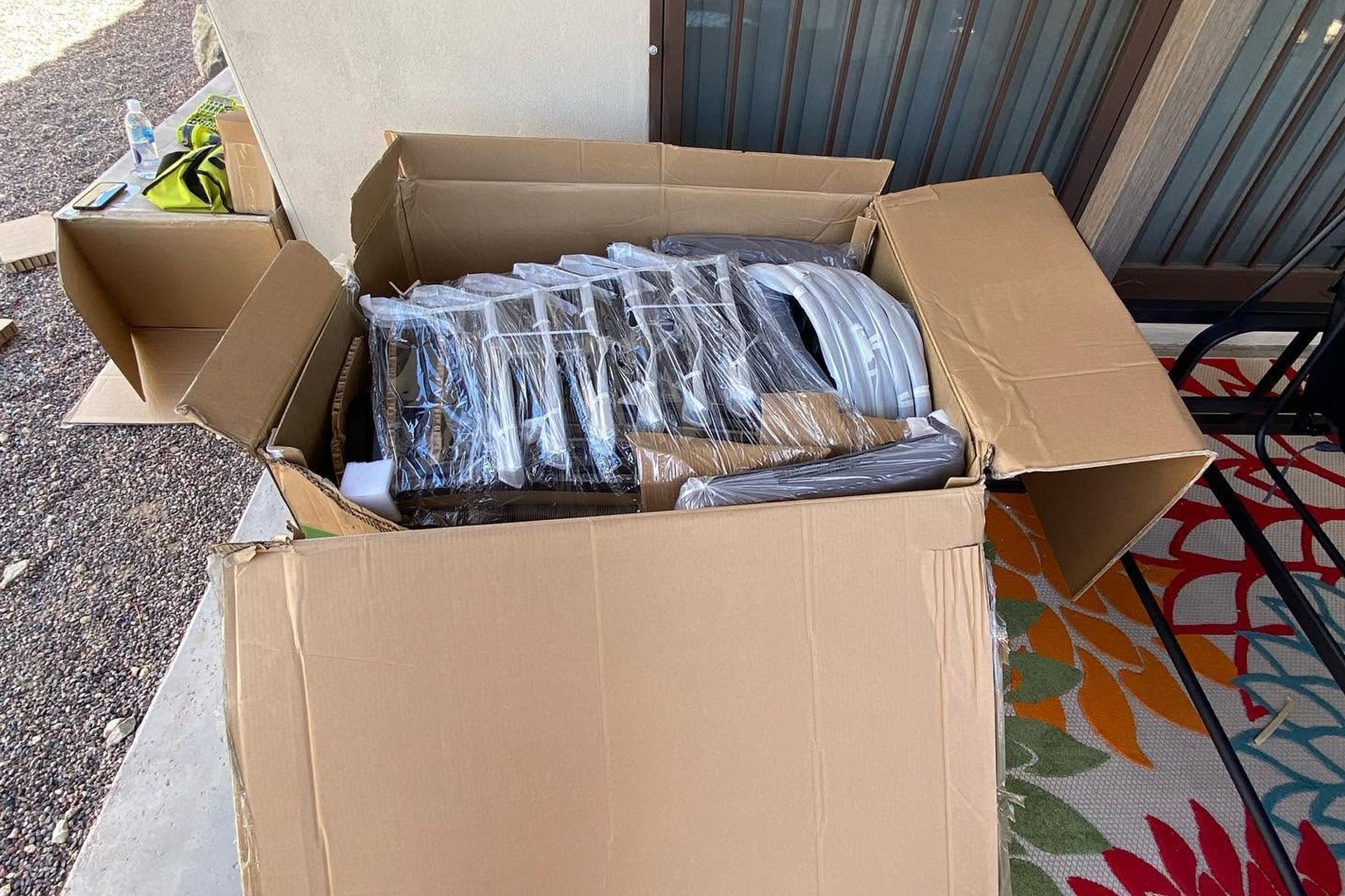 Open cardboard box filled with wrapped black items on a porch beside a colorful rug