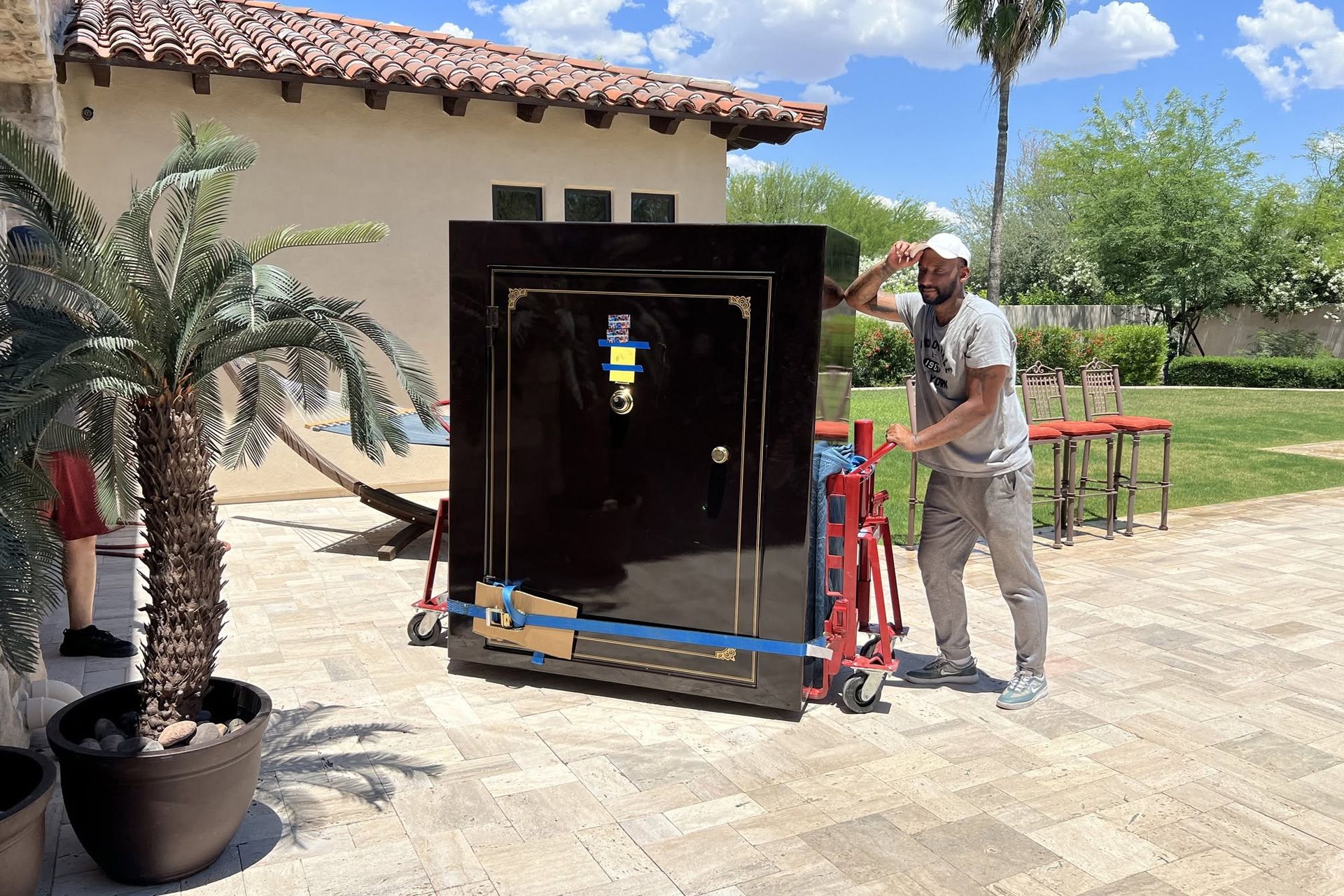 Man moving a large black cabinet on a dolly beside a house patio, with plants and ladder nearby