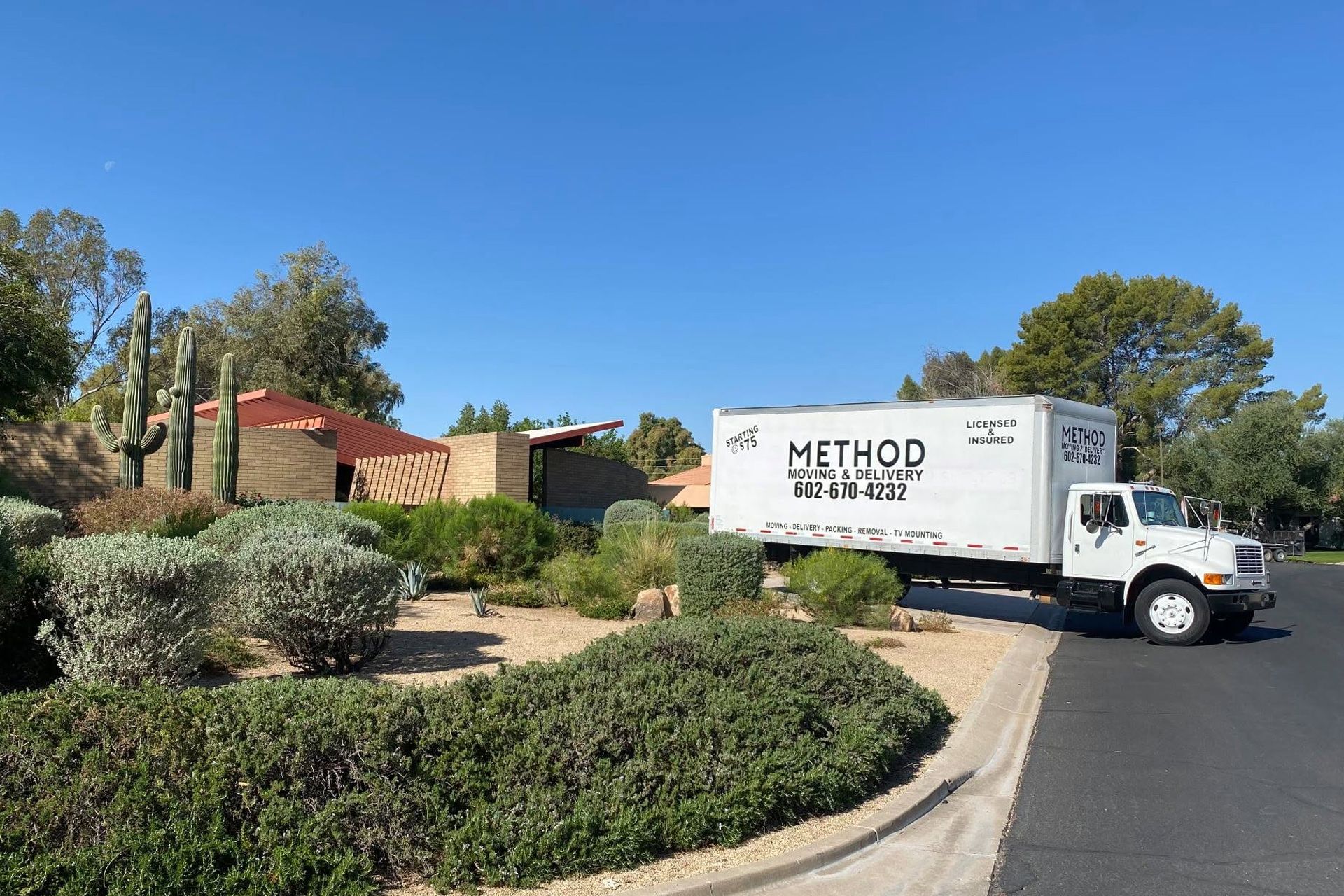 Method Moving truck parked on a curving driveway beside a tan house and landscaped shrubs under a clear blue sky.