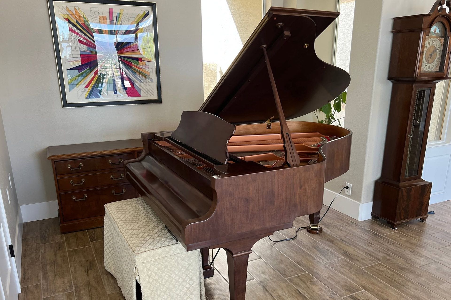 Grand piano in a bright room with framed art, a bench, and a grandfather clock