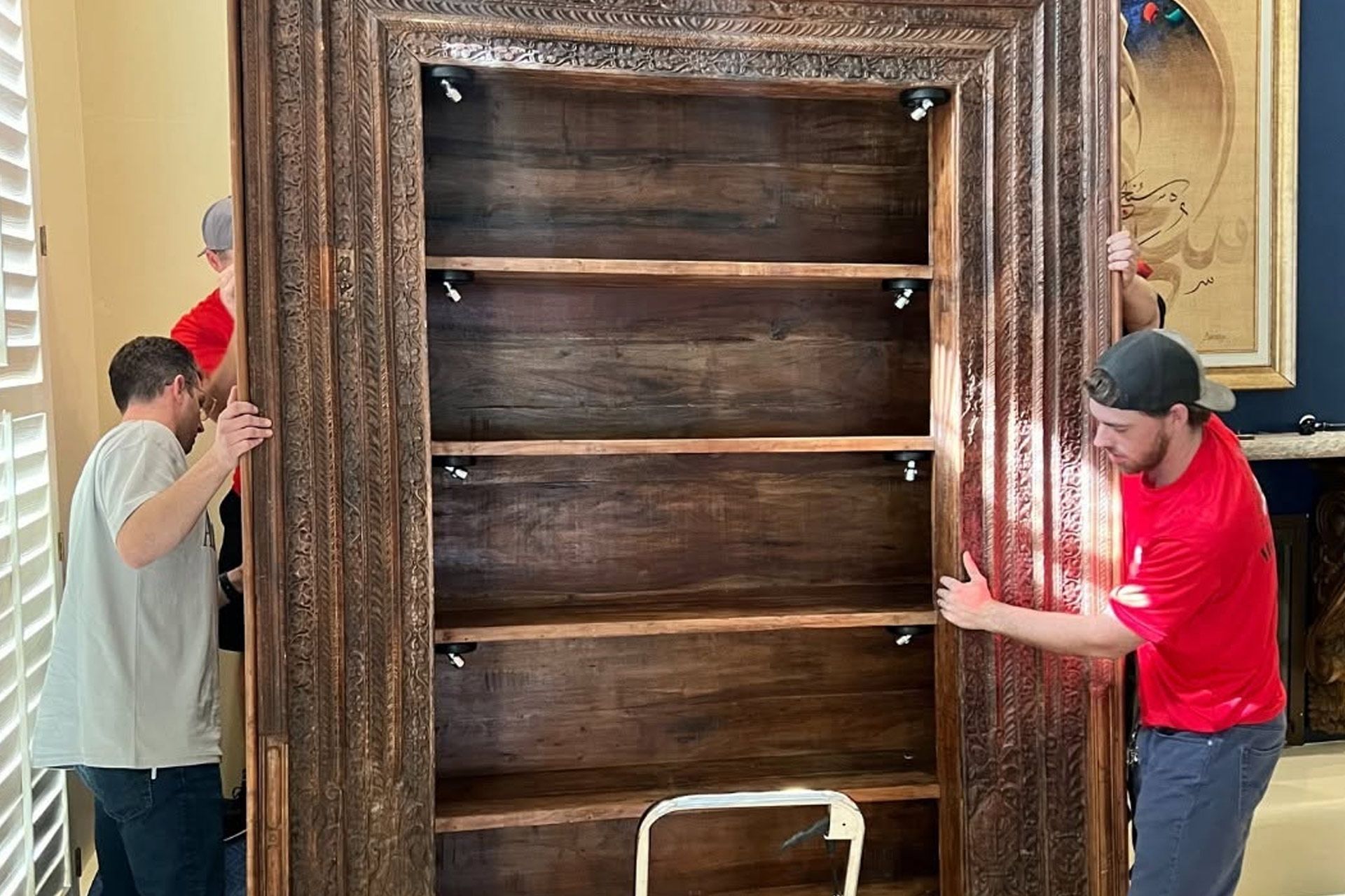 Two people carrying a large wooden cabinet through a doorway, shelves visible inside.
