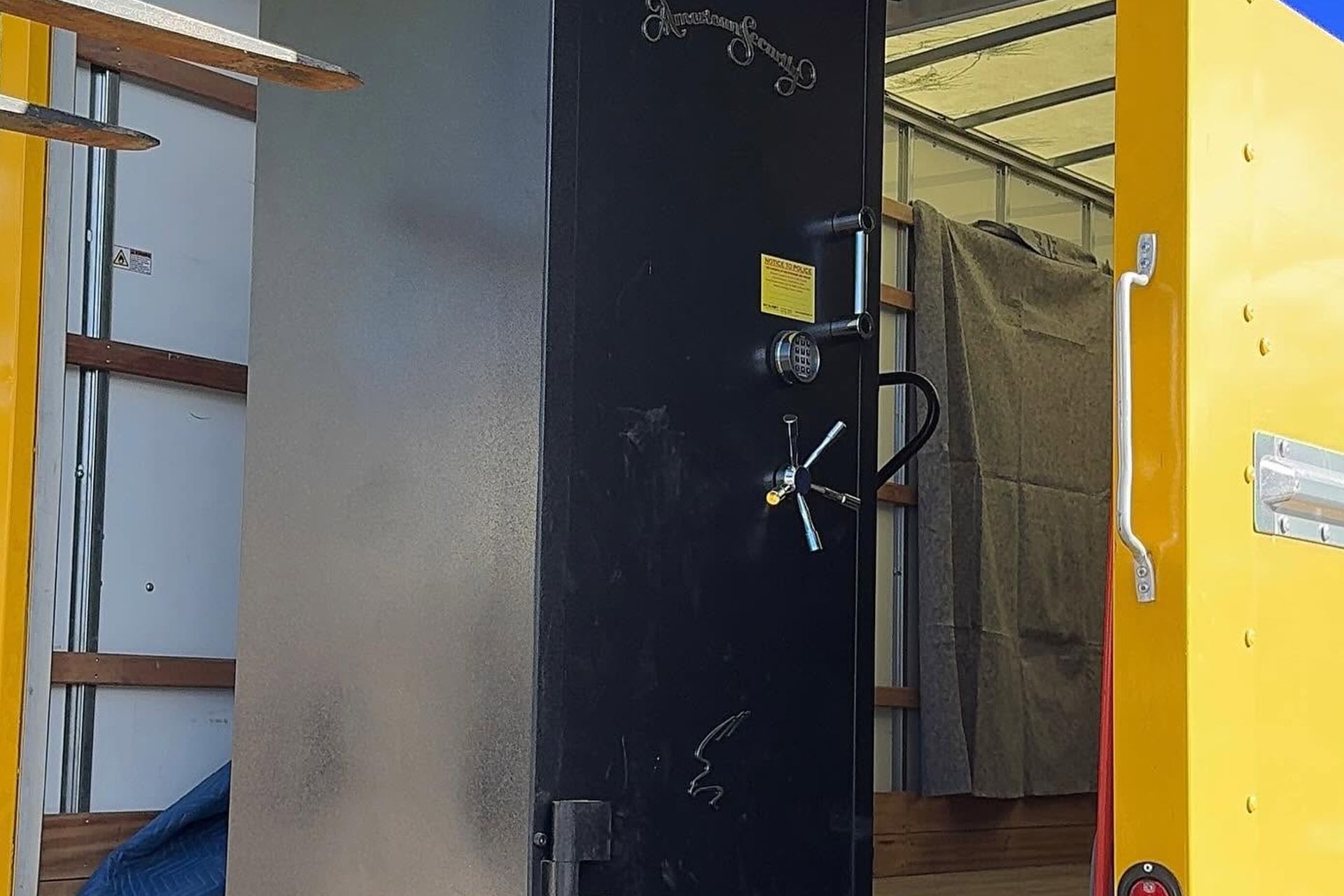 Black safe or vault door in a storage area, with a yellow doorframe and shelving nearby