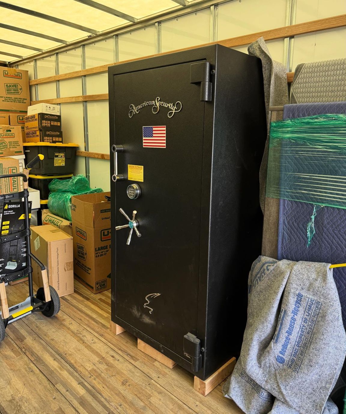 Black safe with American flag sticker in a warehouse, beside stacked boxes and wrapped items.