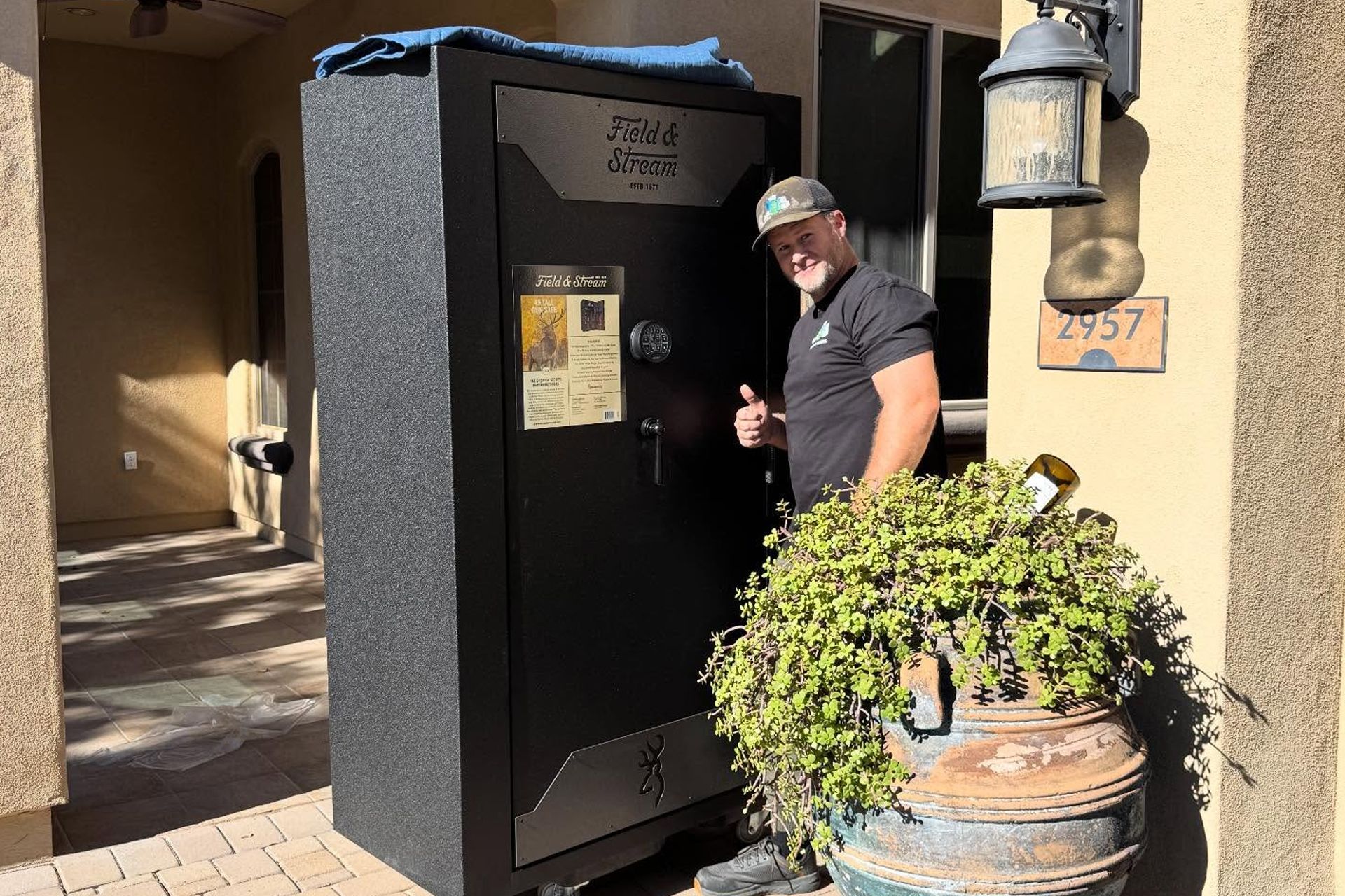 Man standing by a black kiosk outside a building, next to a potted plant and wall lantern.
