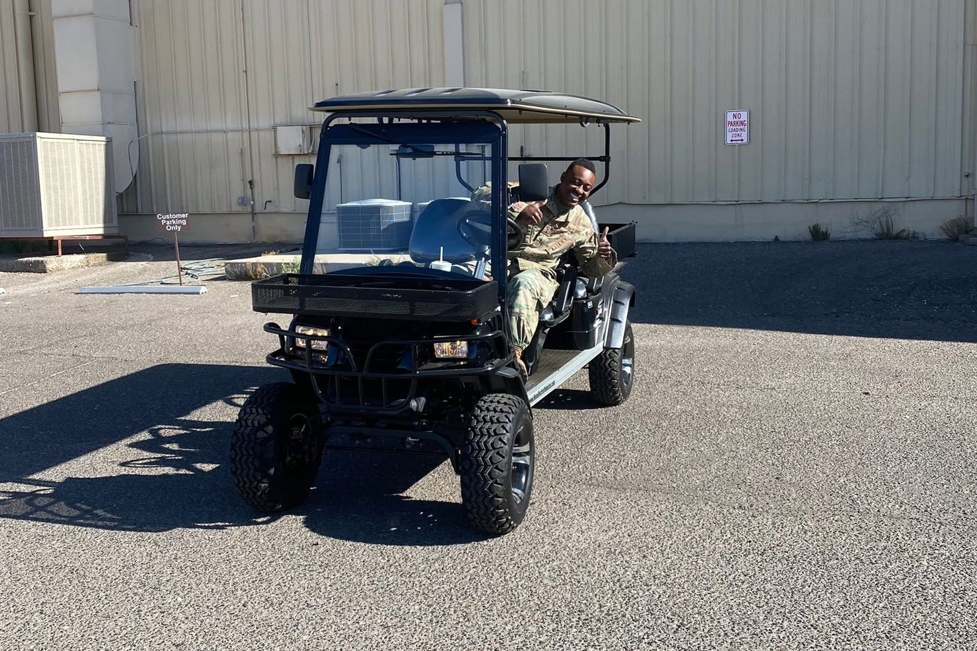 Two people riding in a black utility cart on a gravel lot beside a beige building.