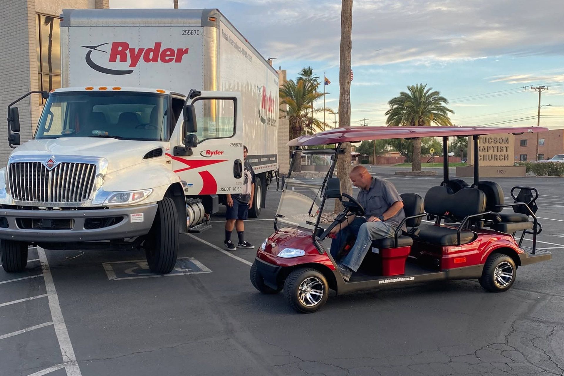 Ryder truck and red golf cart with people in a parking lot at sunset