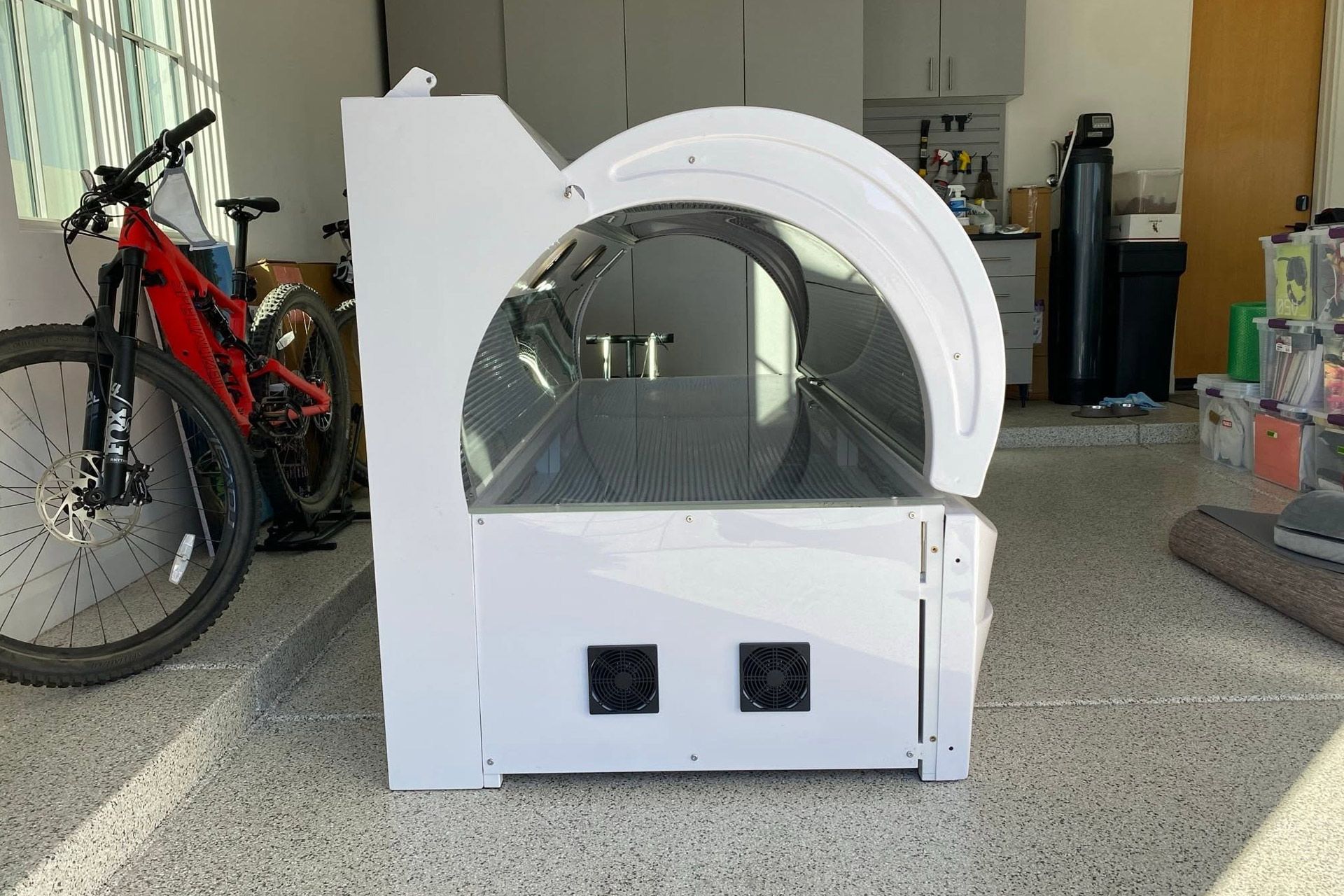 White cylindrical machine with a transparent front panel in a garage, beside a red bicycle and storage items