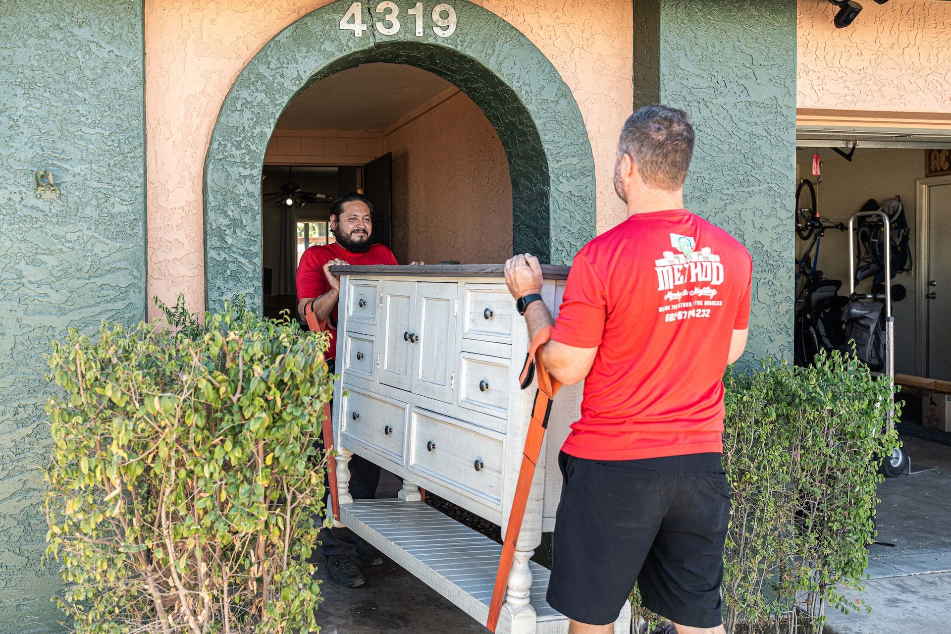 Two movers loading a white cabinet into a house with an arched doorway.