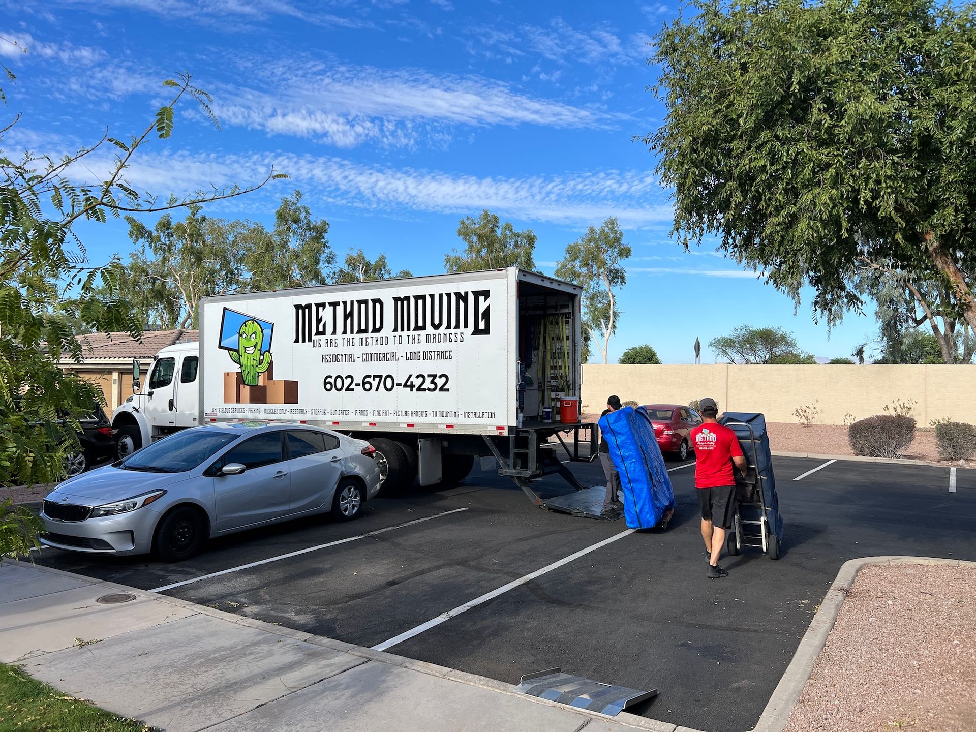 Moving truck and workers loading items beside a silver car in a driveway under a blue sky.