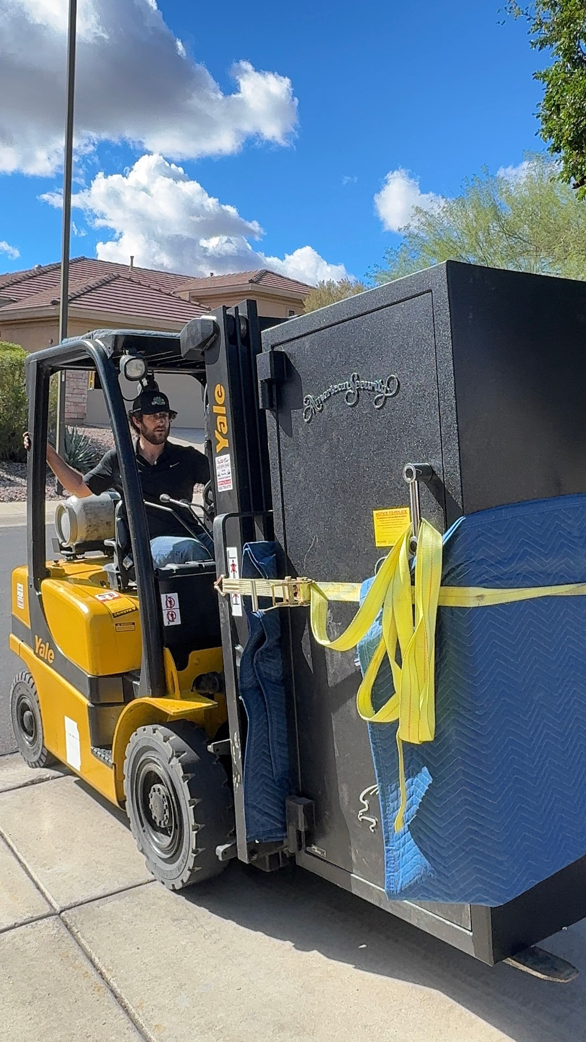 Yellow forklift carrying a large black-and-blue bin outdoors on a sunny day