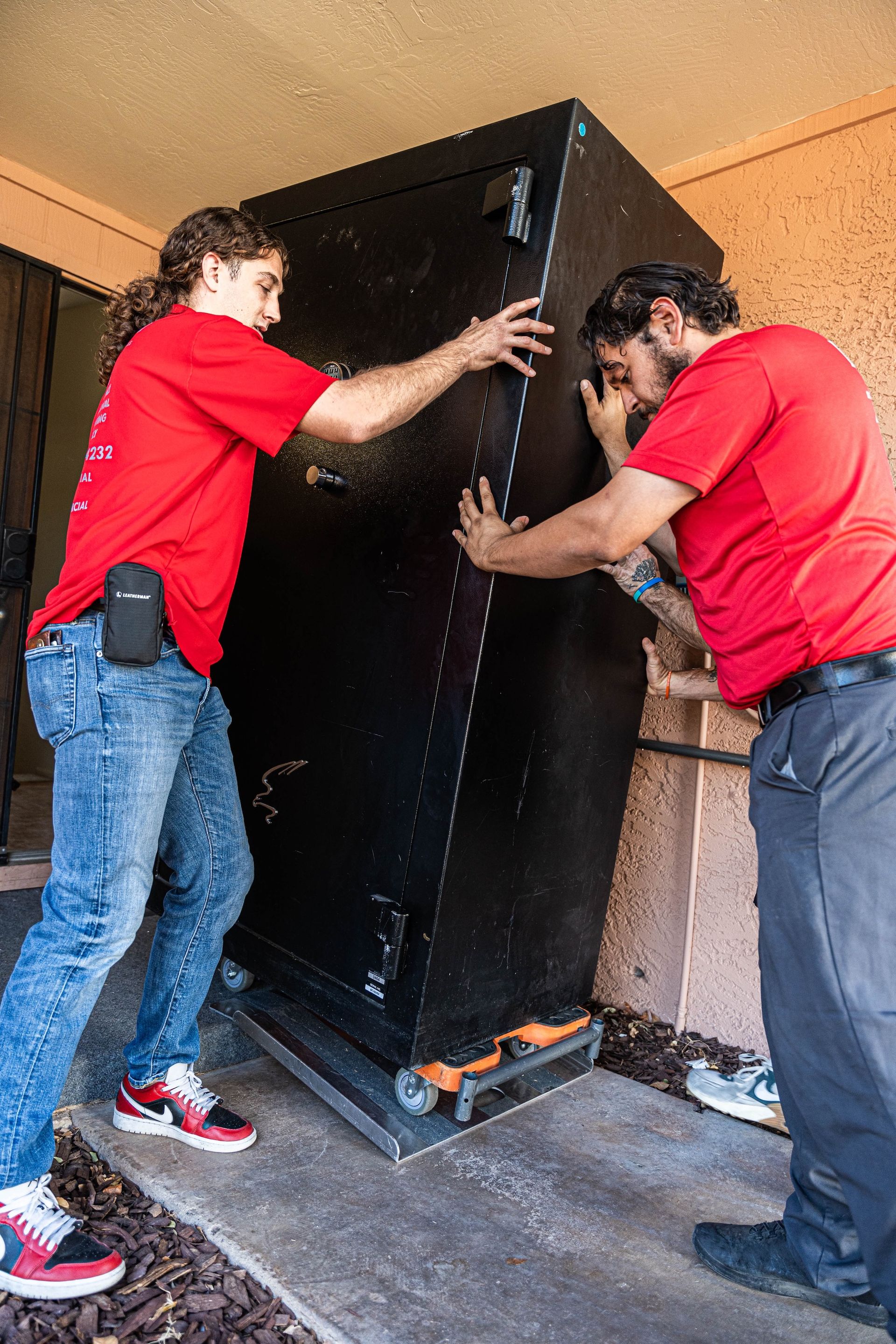 Two workers in red shirts move a large black cabinet through a doorway.