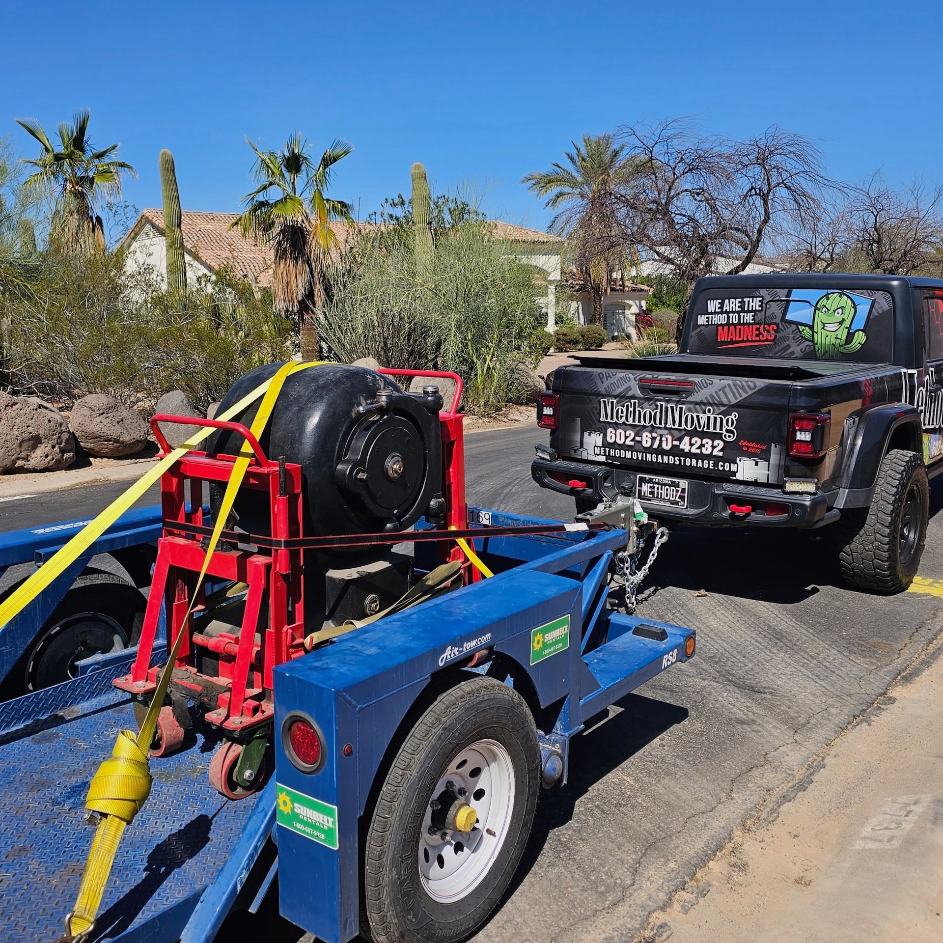 Blue utility trailer with red equipment behind a black pickup truck on a sunny street.