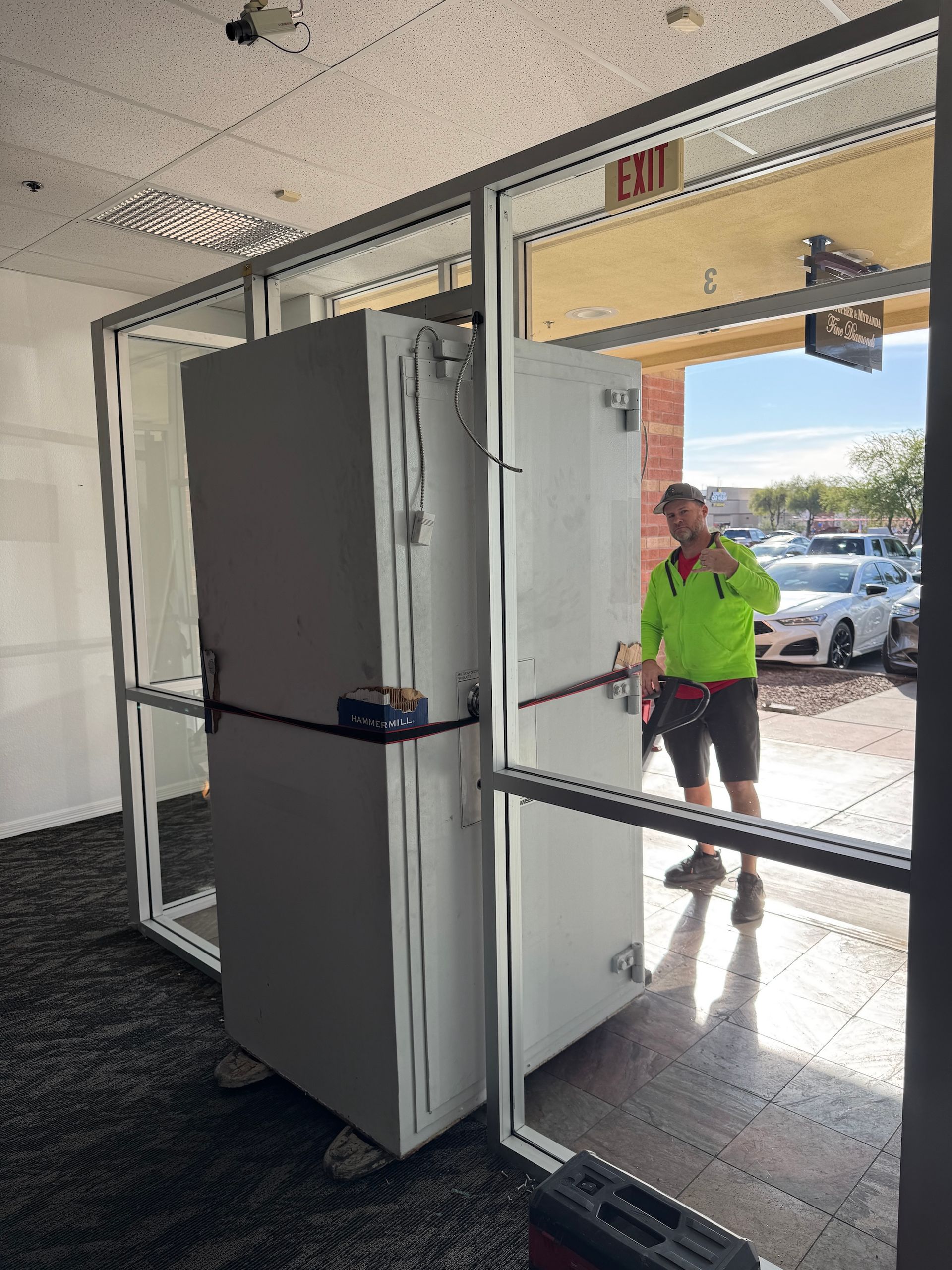 Open glass storefront doors with a large white cabinet being moved inside; person in neon shirt stands outside.