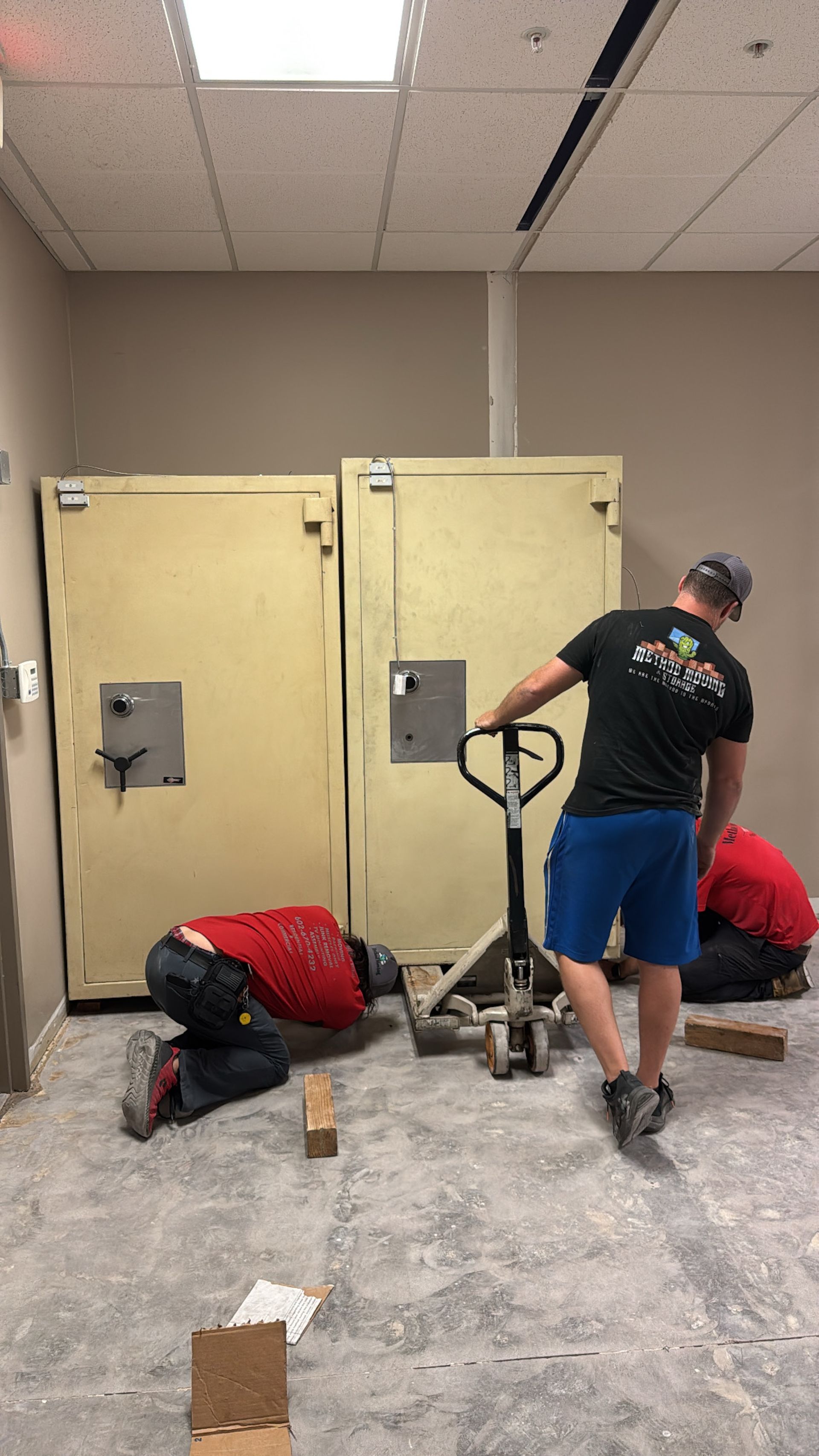 Two workers near large beige doors, one operating a floor machine in a dusty room.