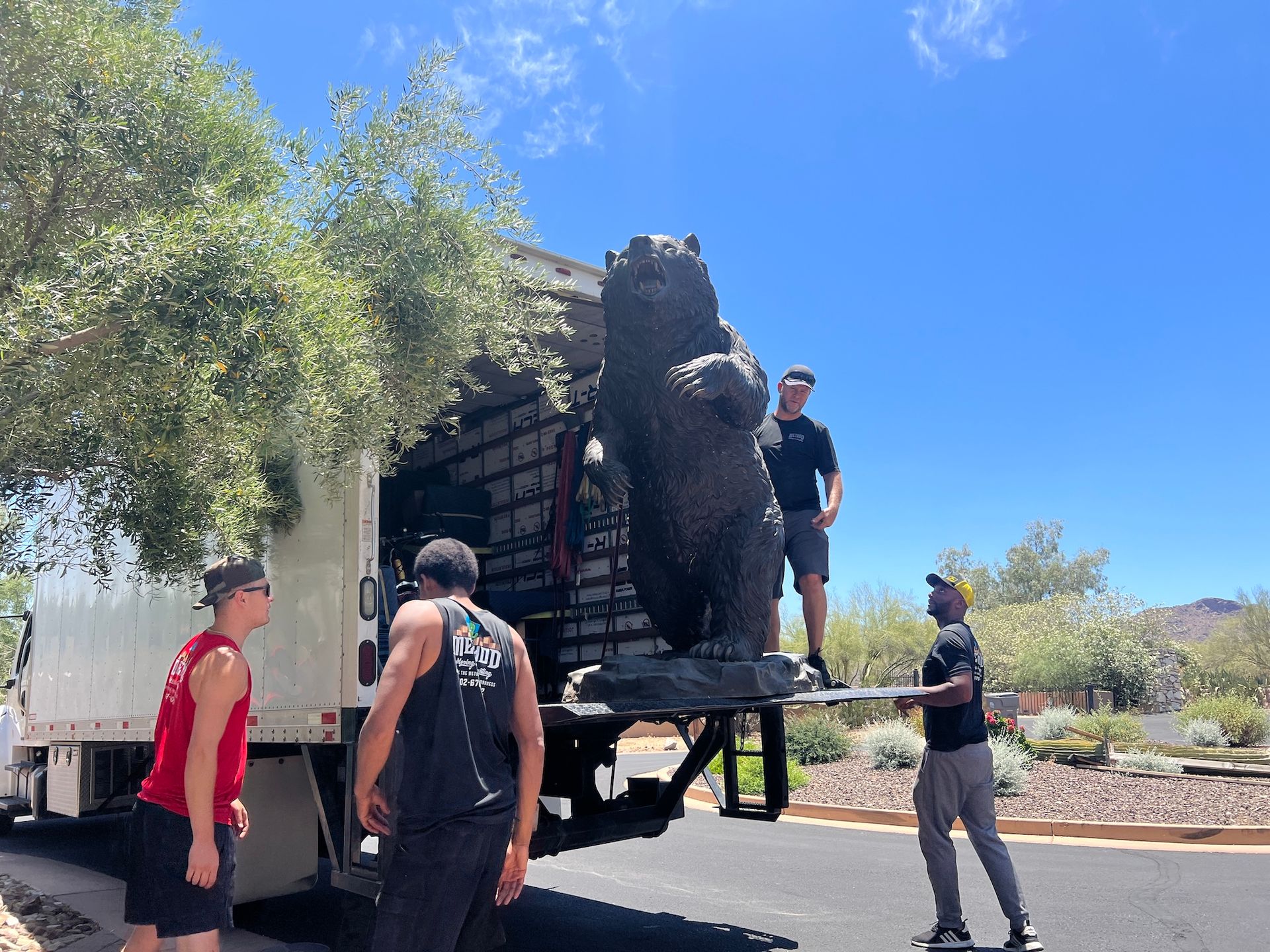 Workers unloading a large black sculpture from a truck in a sunny outdoor area