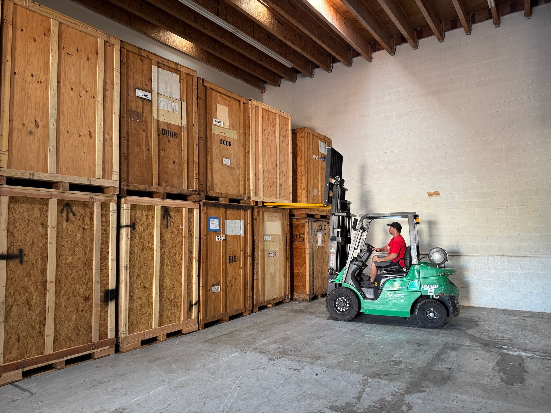 Green forklift in a warehouse aisle beside stacked wooden crates; person in red shirt driving.