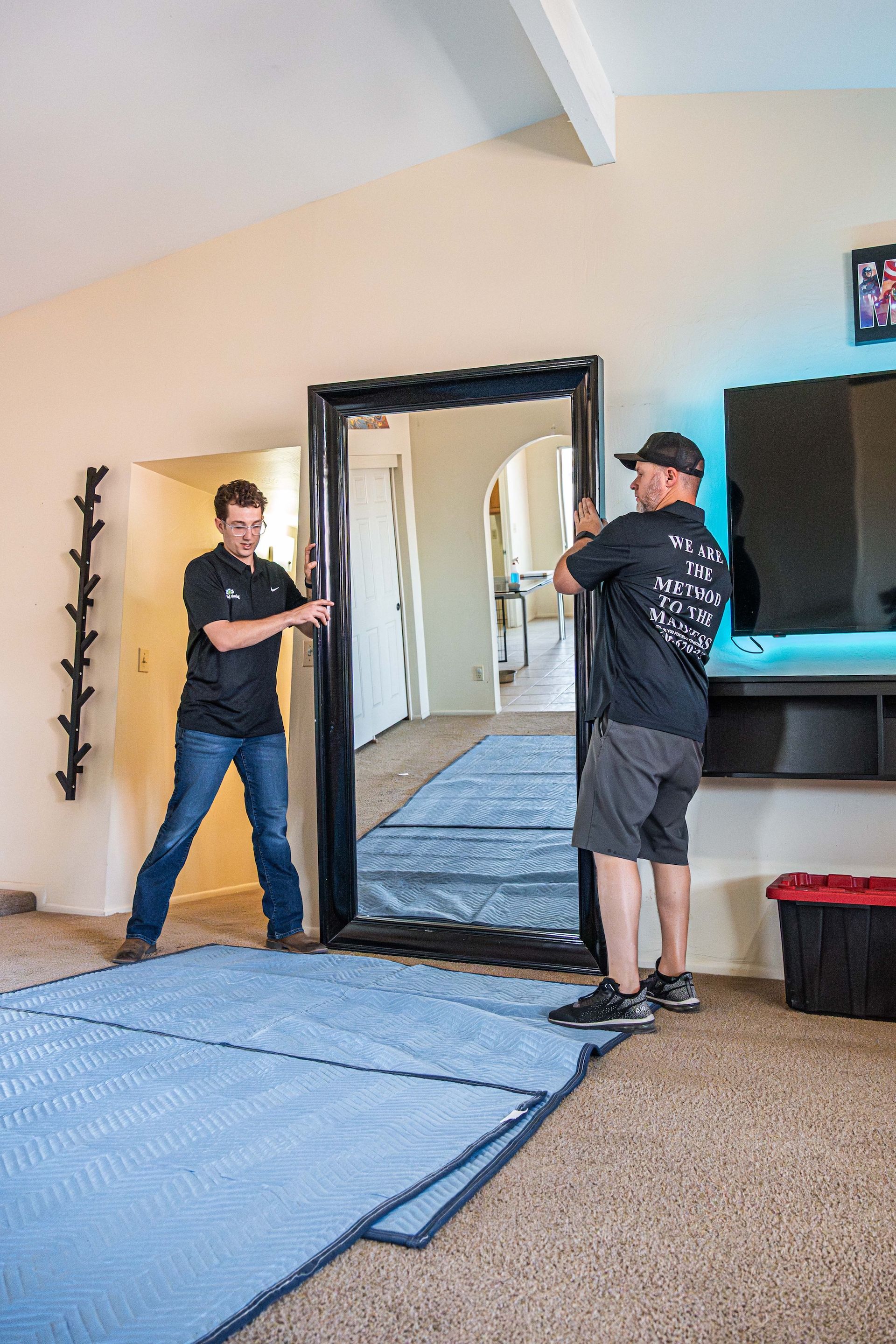 Two people in a living room adjusting a large framed mirror or doorway on a blue floor mat.