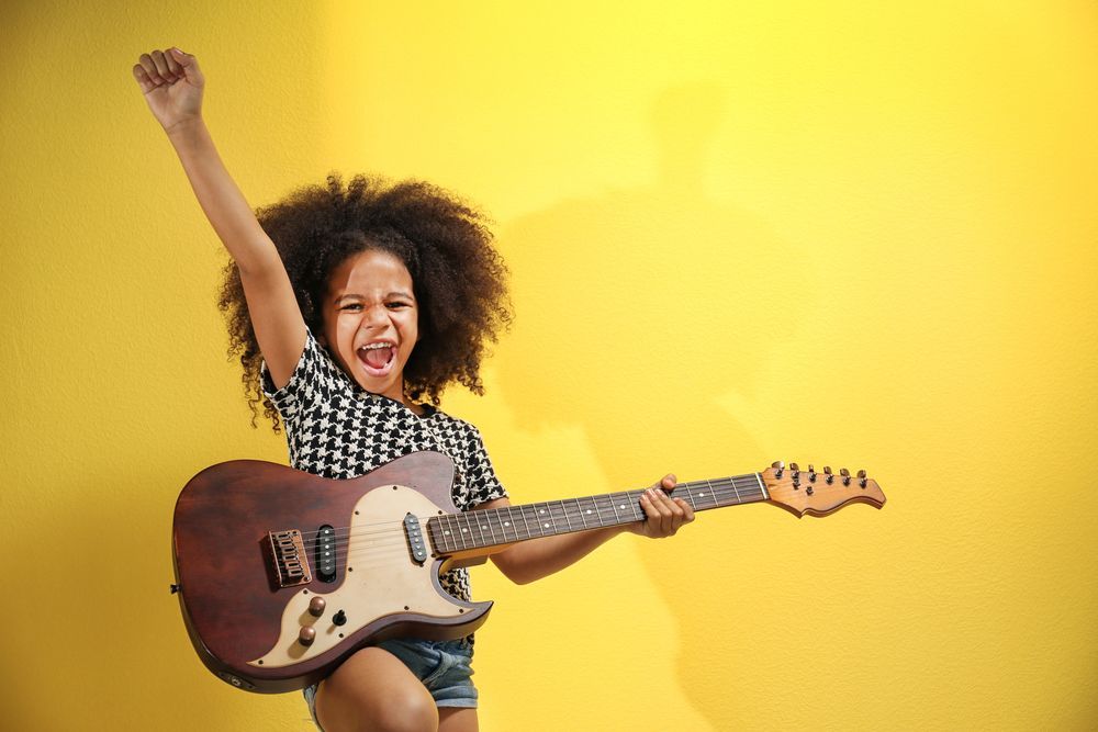 A young girl is playing an electric guitar on a yellow background.