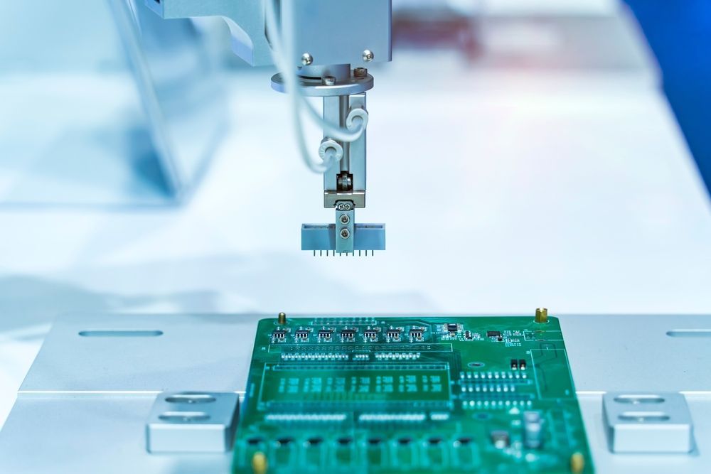 A robotic arm with a specialized nozzle hovers over a green printed circuit board in an industrial setting.