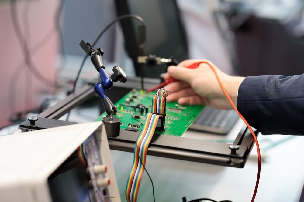 Hands working on a green circuit board with test leads and equipment on a lab bench