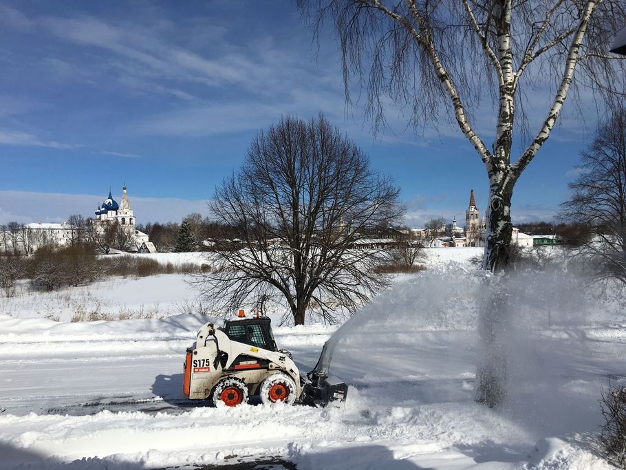 Snowblower clearing a snowy yard on a sunny winter day, with trees and a church in the background