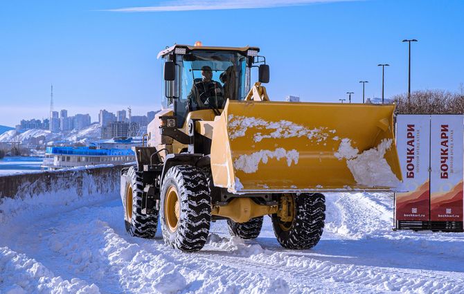 Yellow snowplow clearing a snow-covered road beside a river on a bright winter day