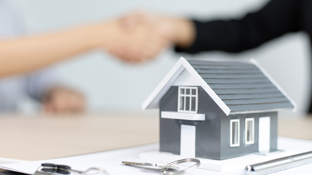Two people are shaking hands in front of a model house and keys.