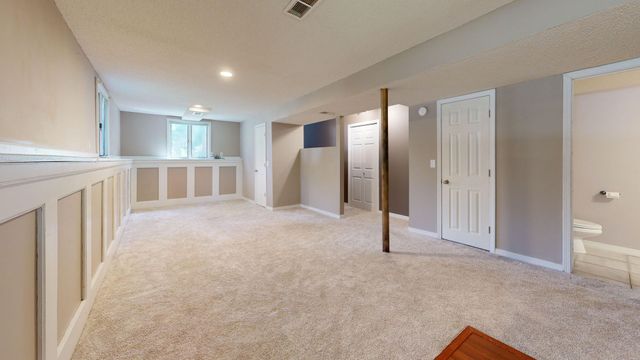 Empty beige carpeted room with white trim, slanted ceiling, doors, and a bright windowed area beyond.