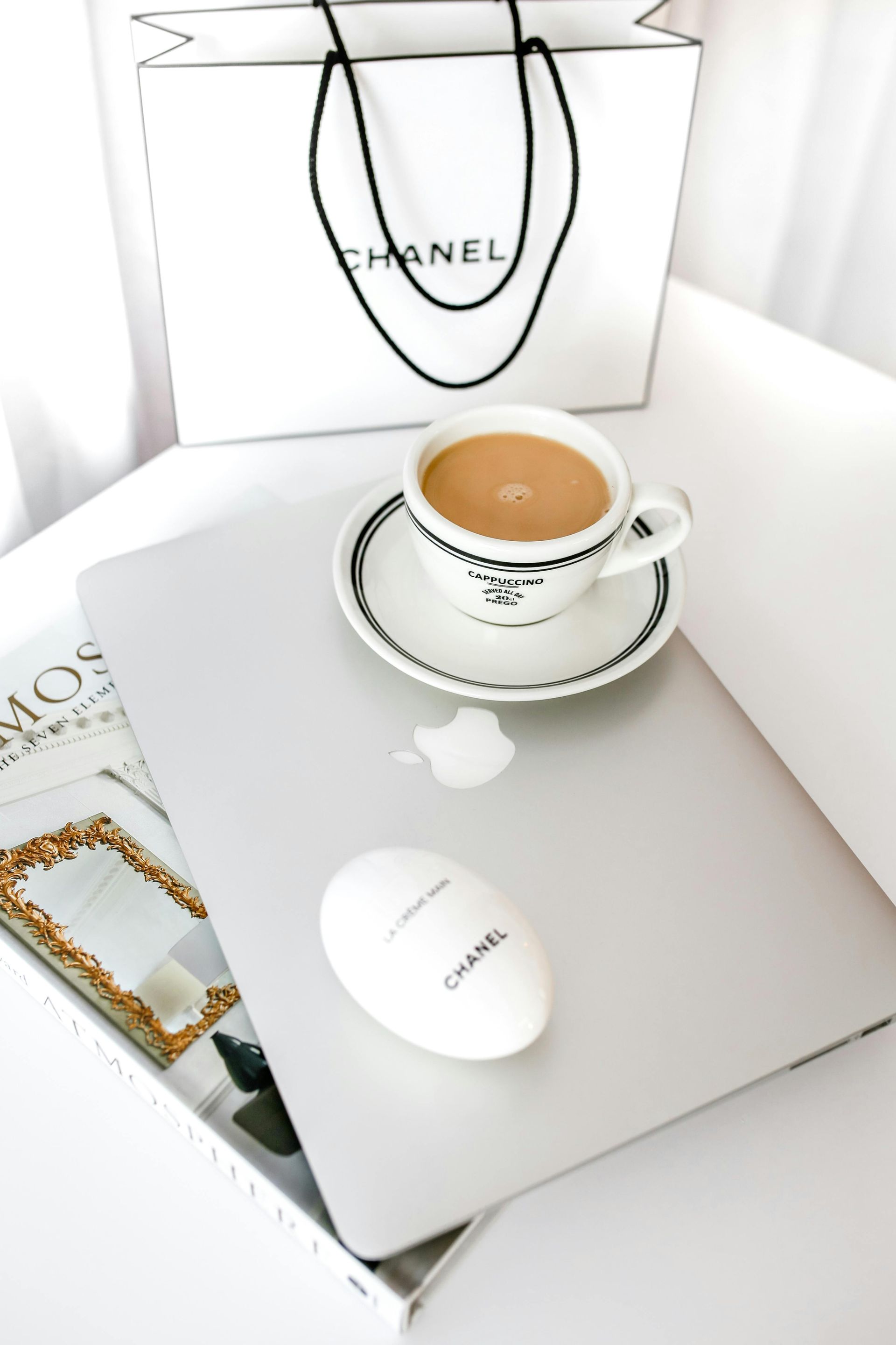 Productos de Chanel exhibidos en una computadora portátil, con una taza de café y una bolsa de compras sobre una superficie blanca.