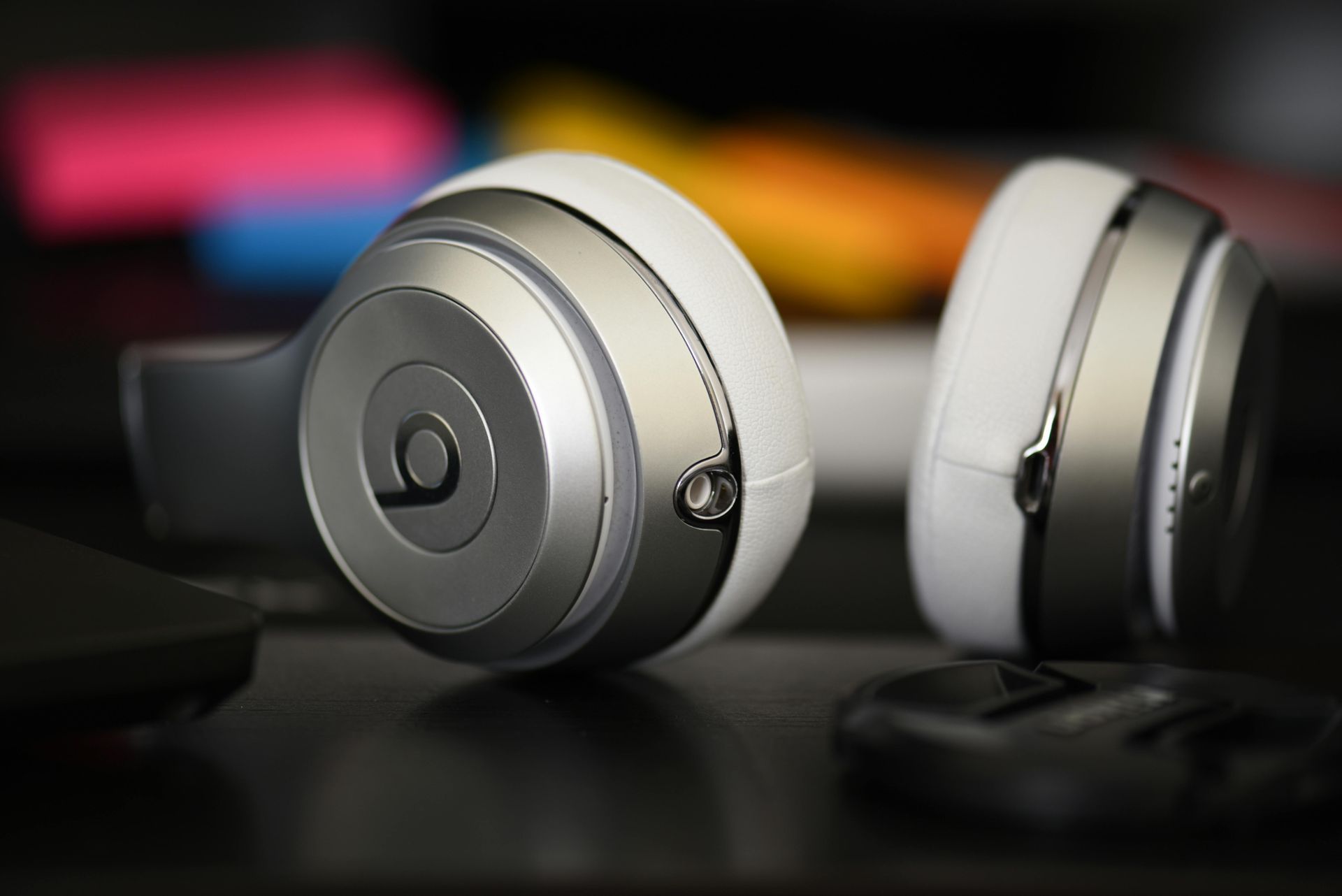 Auriculares Beats blancos y plateados sobre una superficie oscura y reflectante, con fondo colorido borroso.