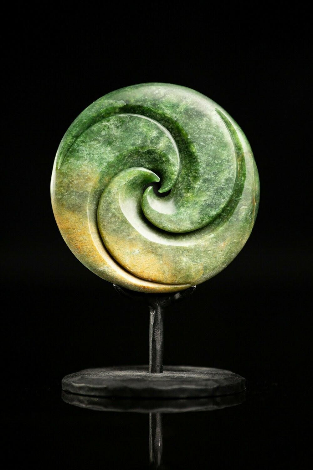 A green and yellow sculpture of a pounamu koru spiral on a stand with a black background.