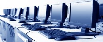 Row of computer monitors, keyboards, and mice on a white desk in a classroom setting.