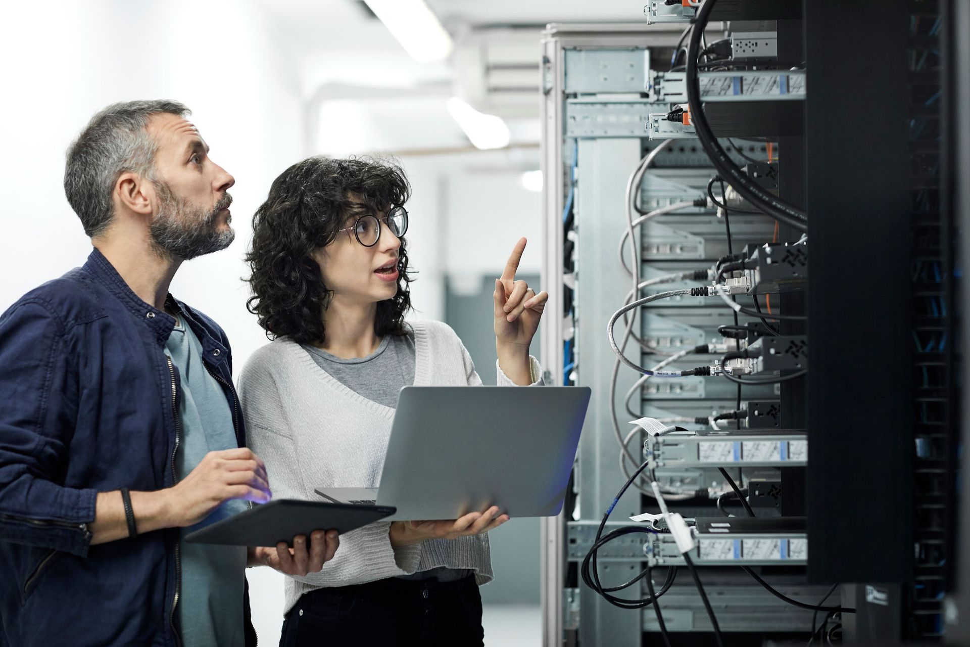 A female technician guiding a male engineer with a digital tablet in a server room.