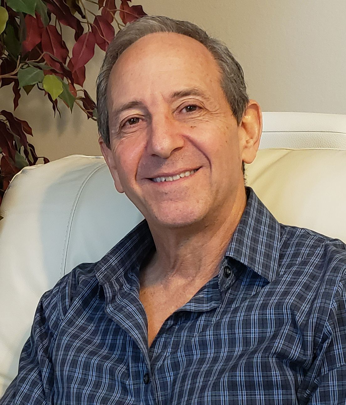 Smiling older man in blue plaid shirt, sitting on a white couch indoors.