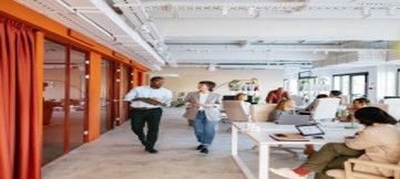 Two people walking in an office with open workspace. The office has white desks, orange curtains.