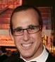 Man with glasses smiling, wearing a dark suit and white dress shirt, at an event.