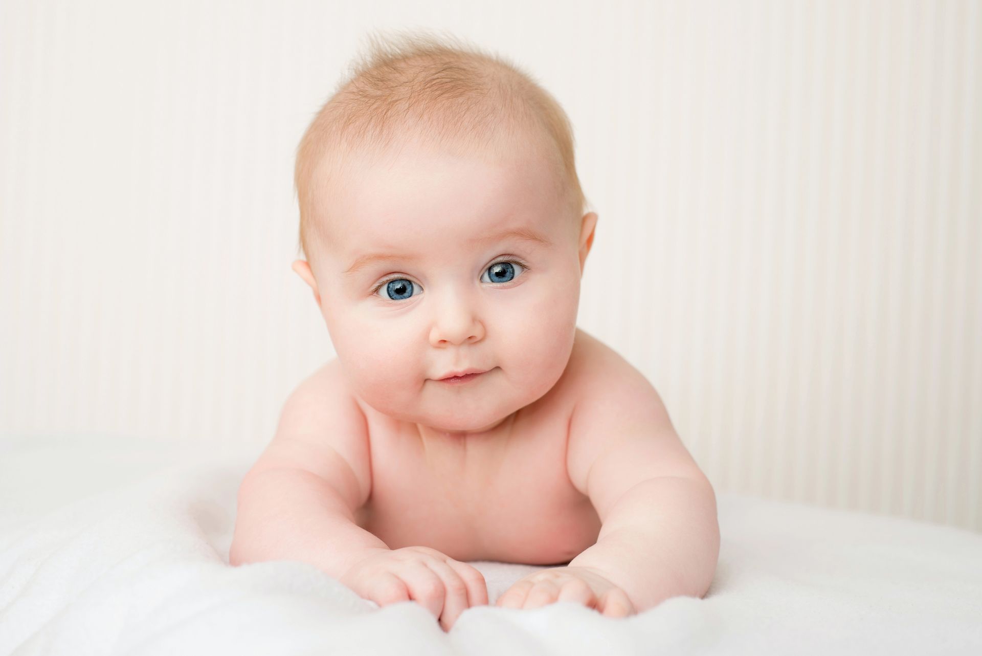 Bambino con pelle chiara e occhi azzurri, sdraiato a pancia in giù su una superficie bianca, con lo sguardo rivolto in avanti.