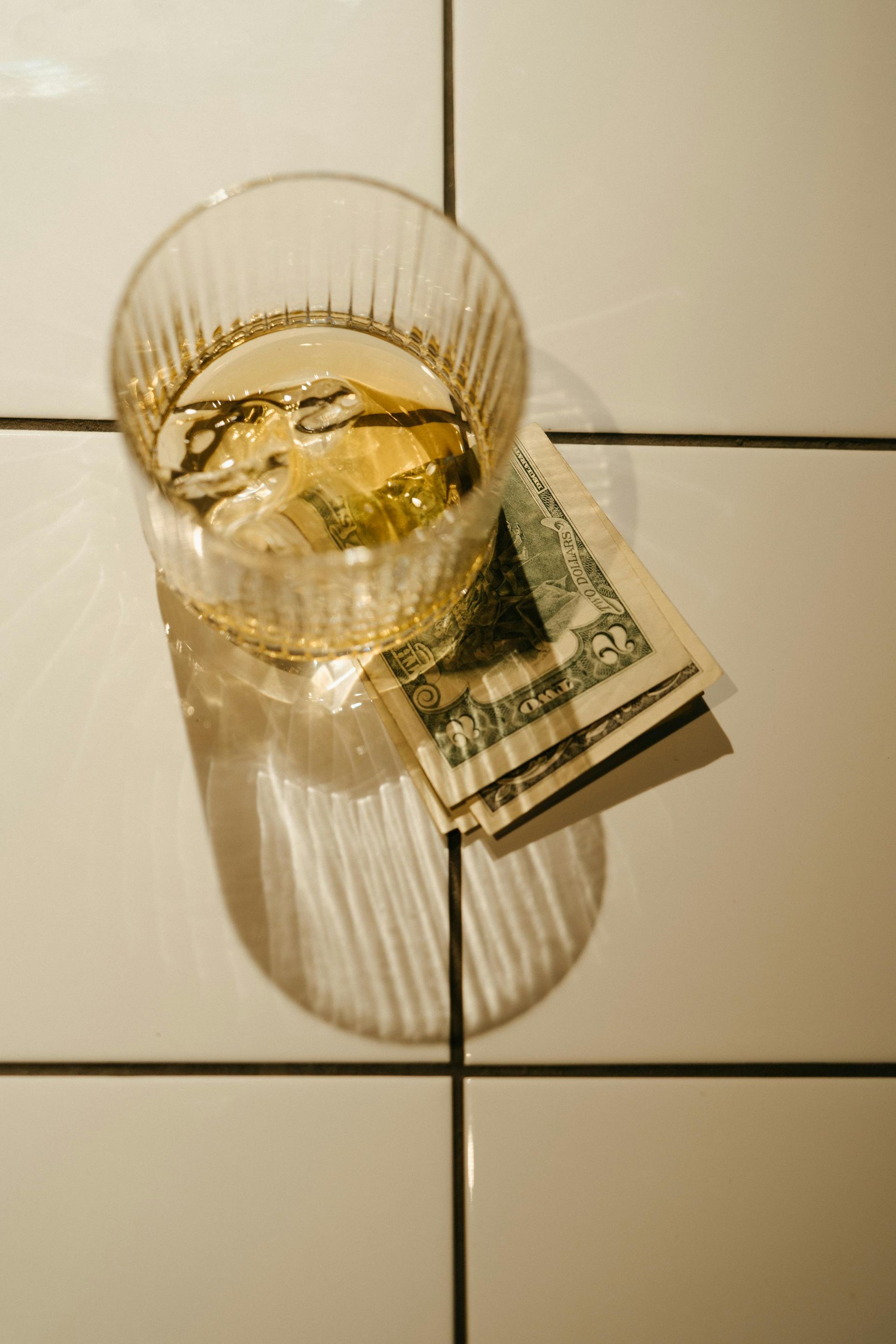 A glass filled with amber liquid and ice sitting on a folded five-dollar bill atop a white tiled surface.