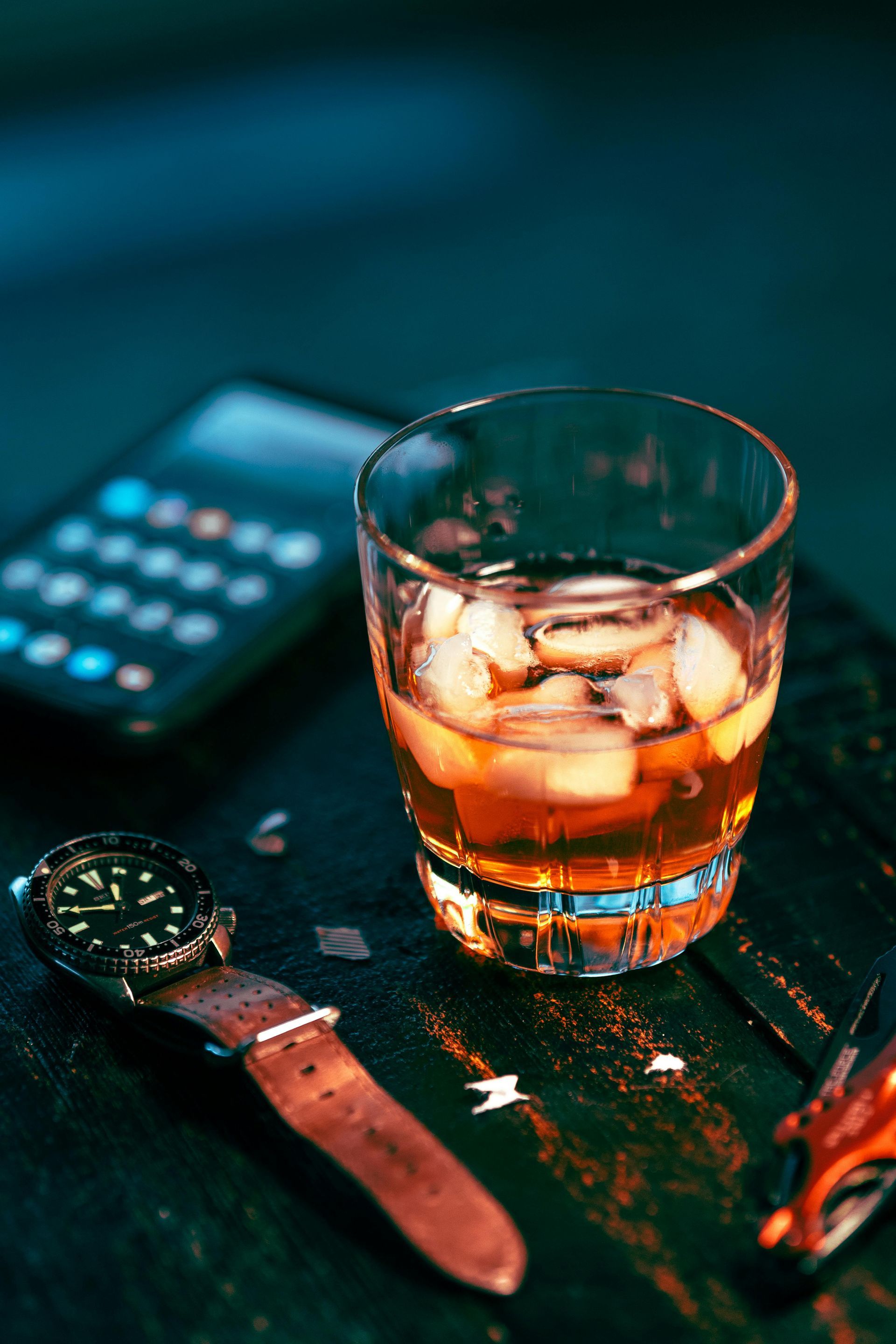 A whiskey glass with ice cubes, a wristwatch with a brown leather strap, and a smartphone on a dark textured surface.