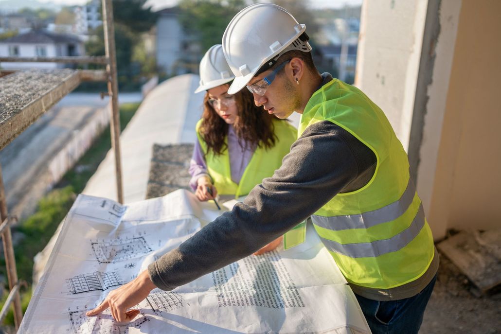 Two construction workers in hard hats review blueprints on a rooftop.