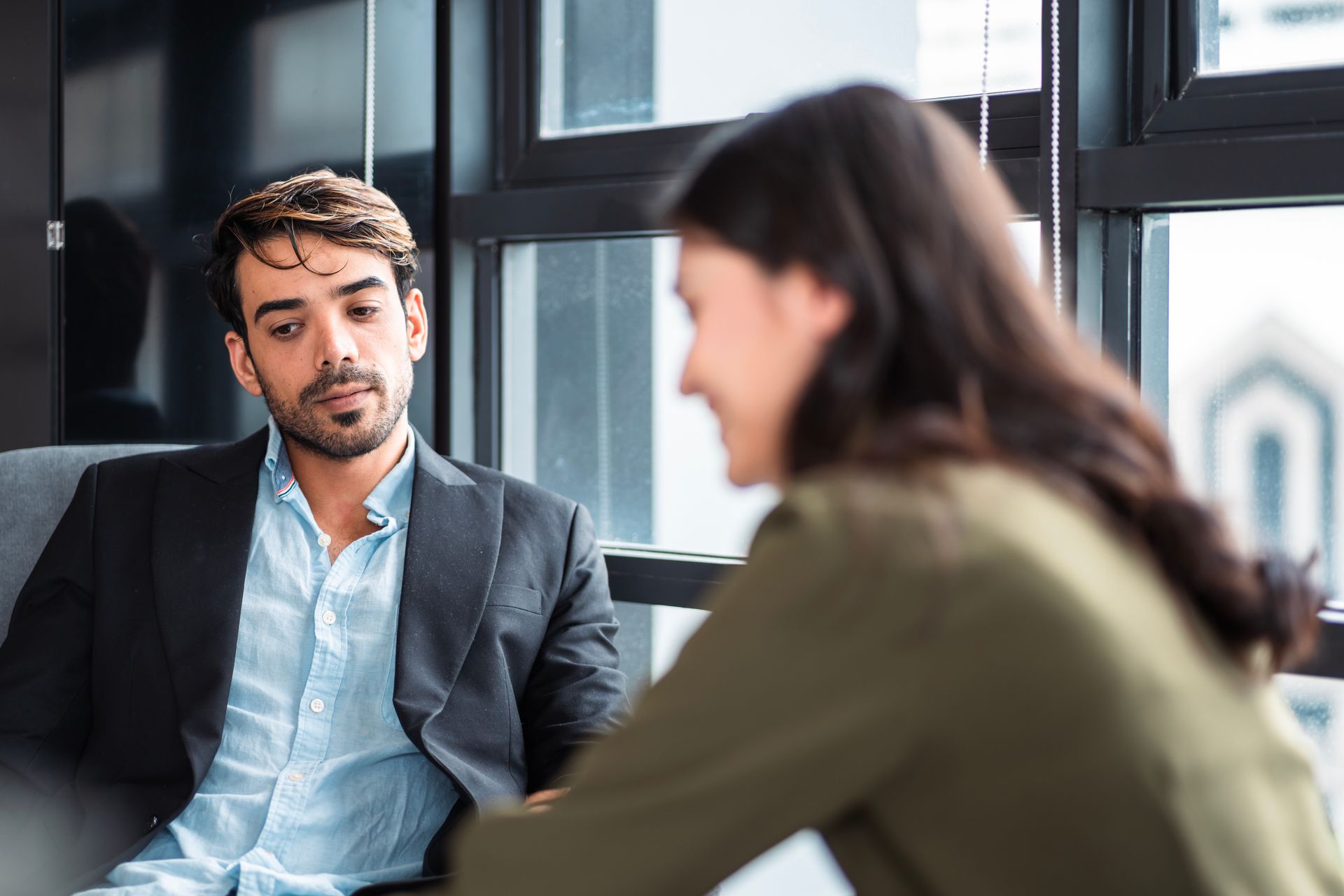 Two people in a modern office having a focused conversation, with one man listening on a sofa.