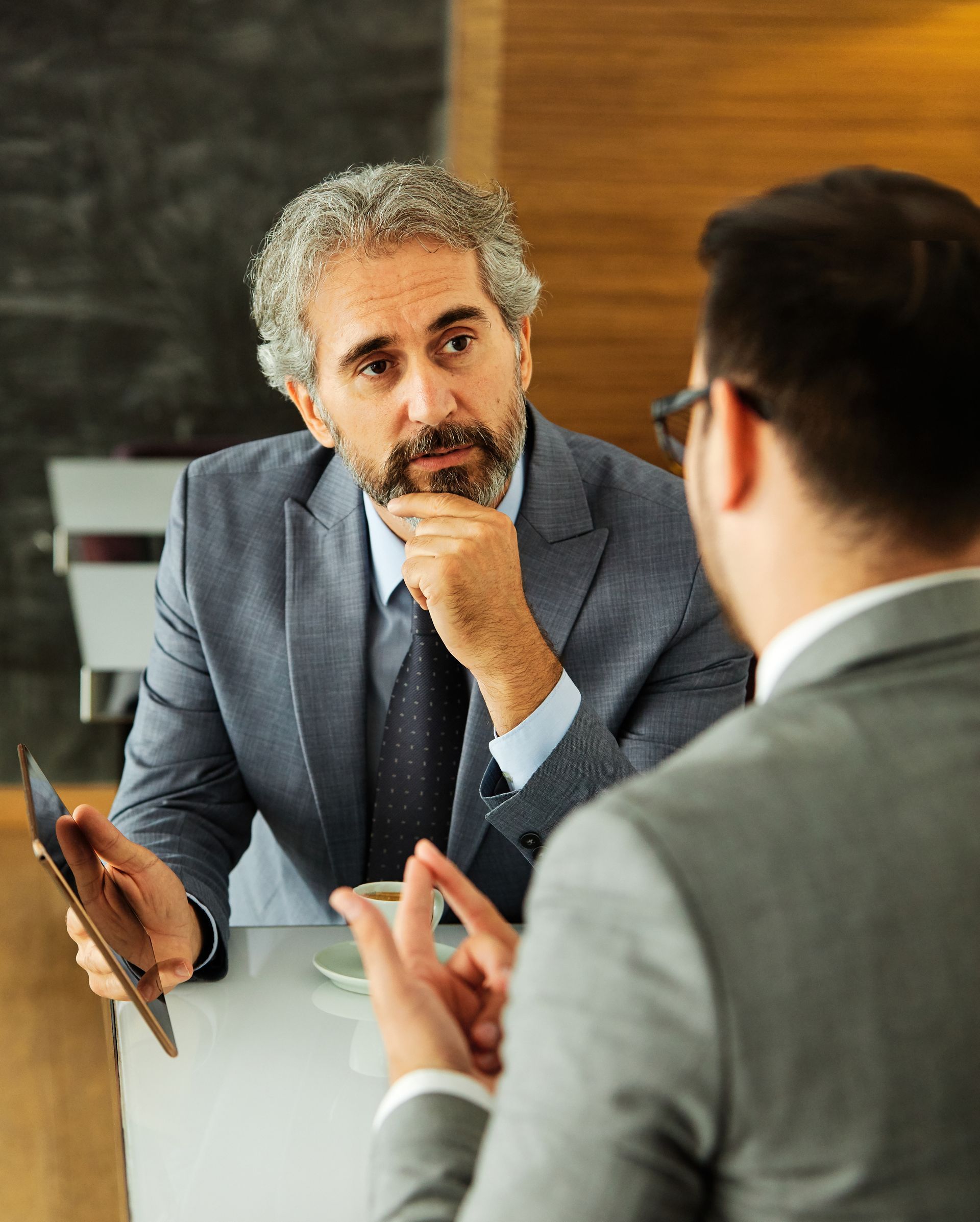 Two coworkers in suits discuss a document in an office, one holding a tablet and listening intently
