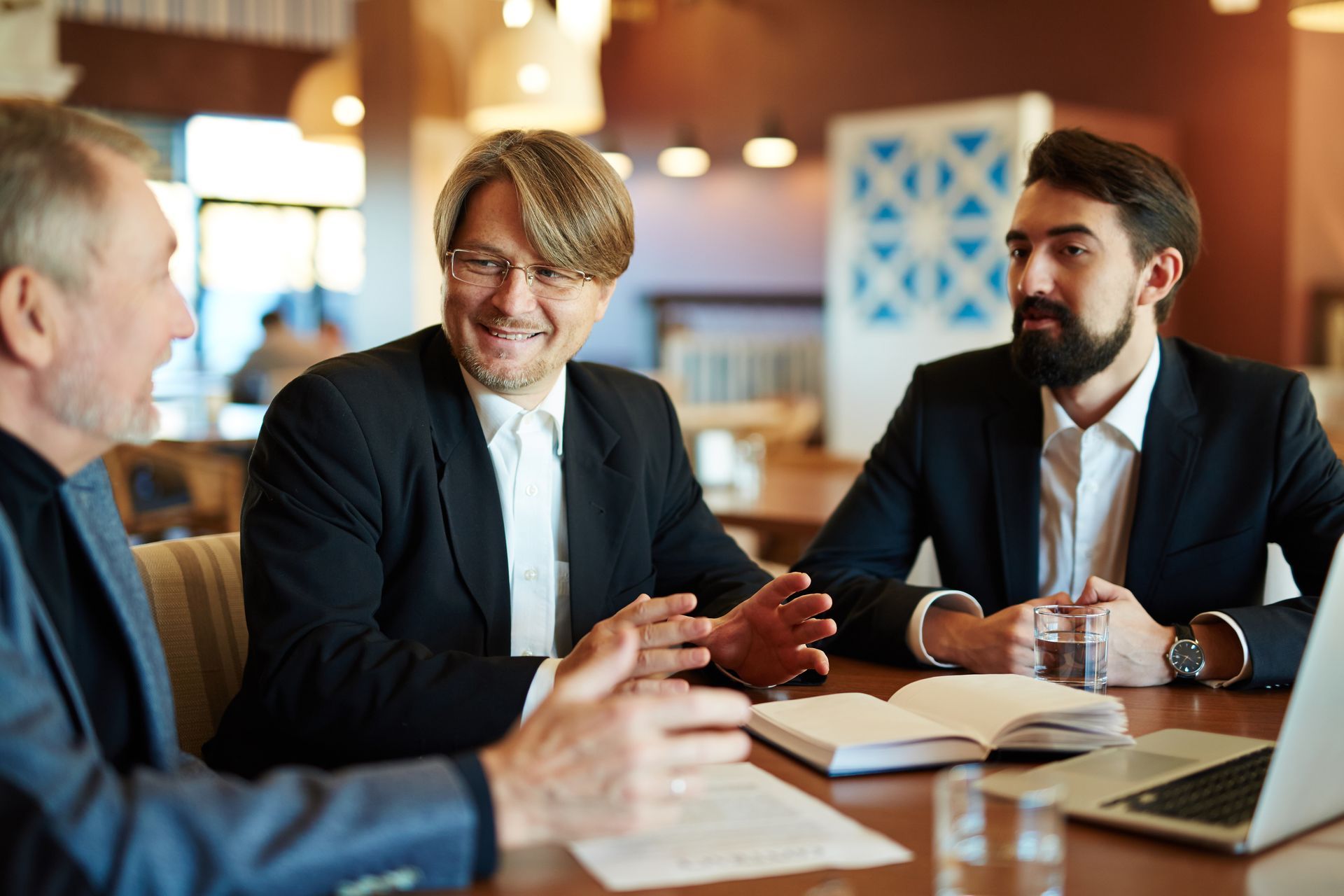 Three people in suits discuss over coffee at a table in a warm indoor meeting space.