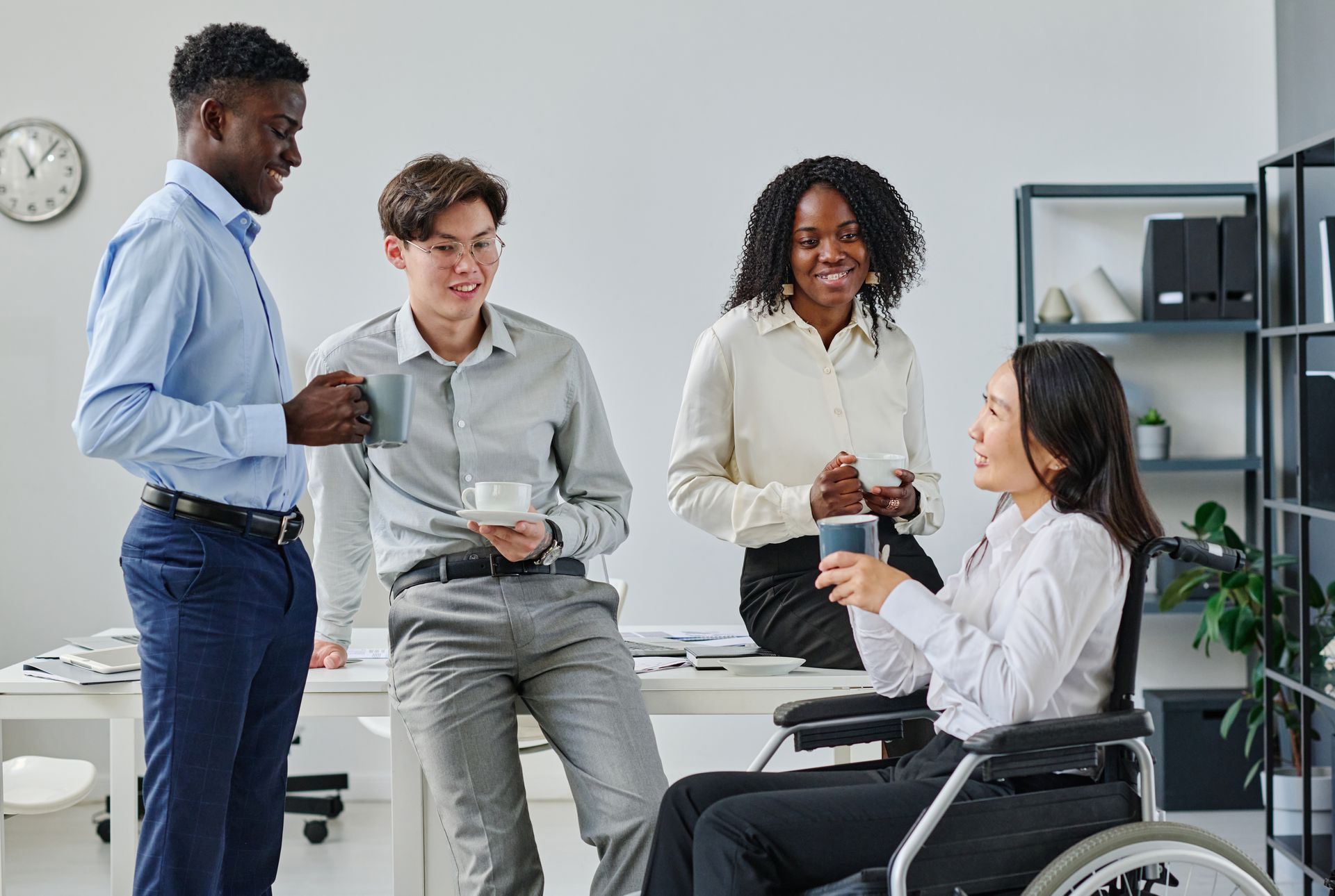 Four coworkers chatting in a bright office, one seated in a wheelchair holding a cup.
