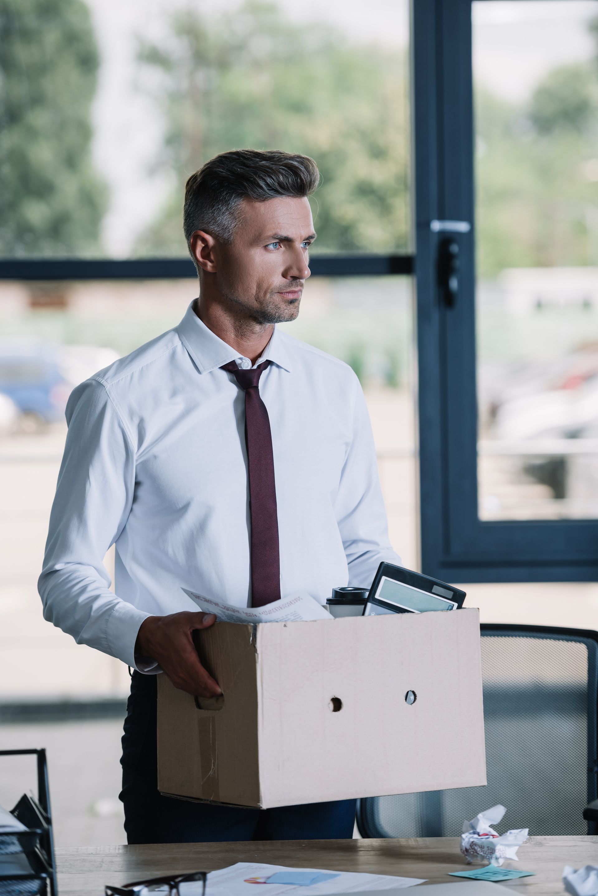 Man holding a cardboard box in an office, wearing a white shirt and tie, looking out a window.