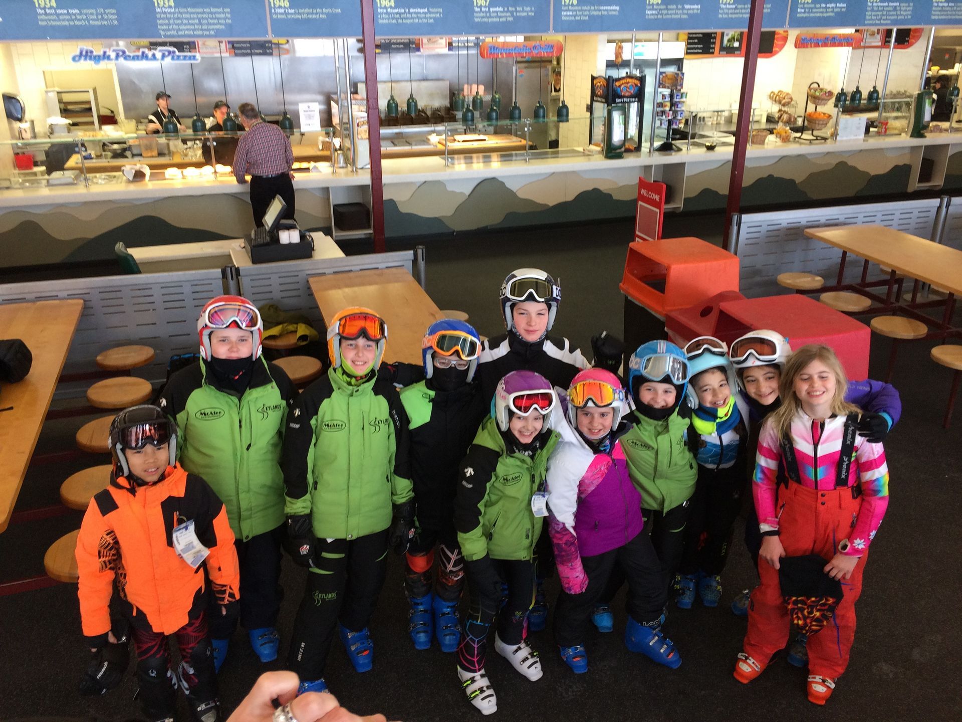 Group of children in ski gear posing, possibly at a ski lodge with a food counter visible.